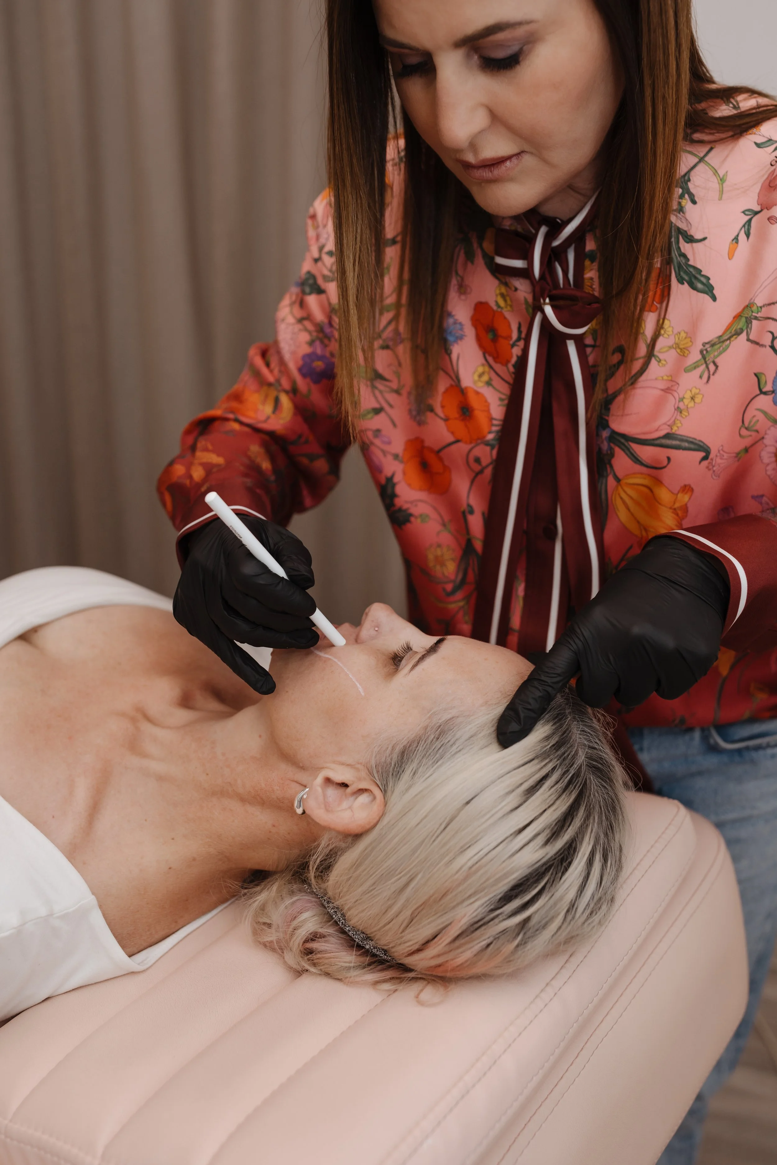 A woman with white hair lying on a cushioned surface receiving a cosmetic treatment on her face from a practitioner wearing black gloves, in a clinical setting.