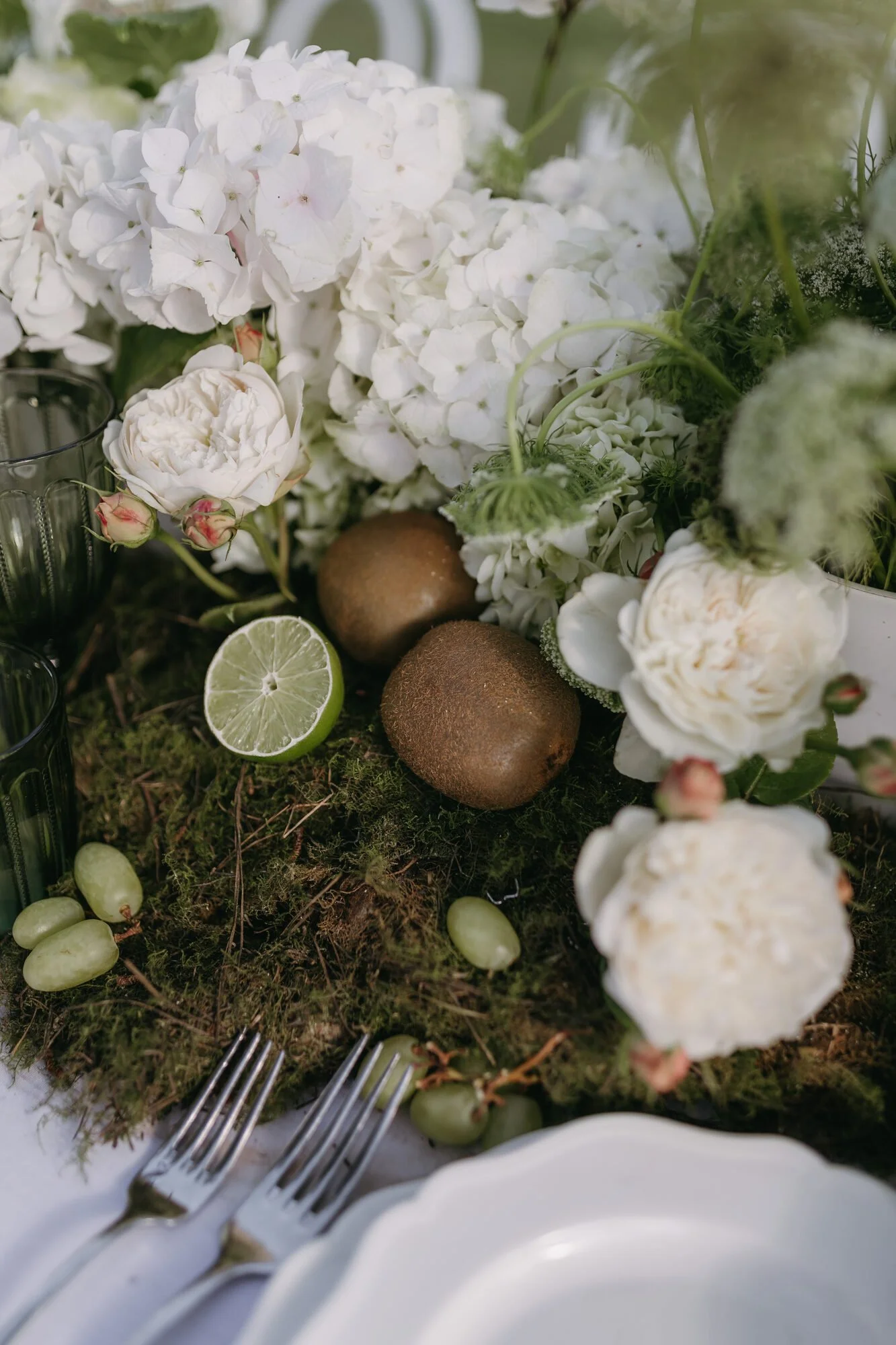 A table centerpiece of white flowers, including hydrangeas and roses, with limes, green grapes, and kiwis, surrounded by silver cutlery and white plates.