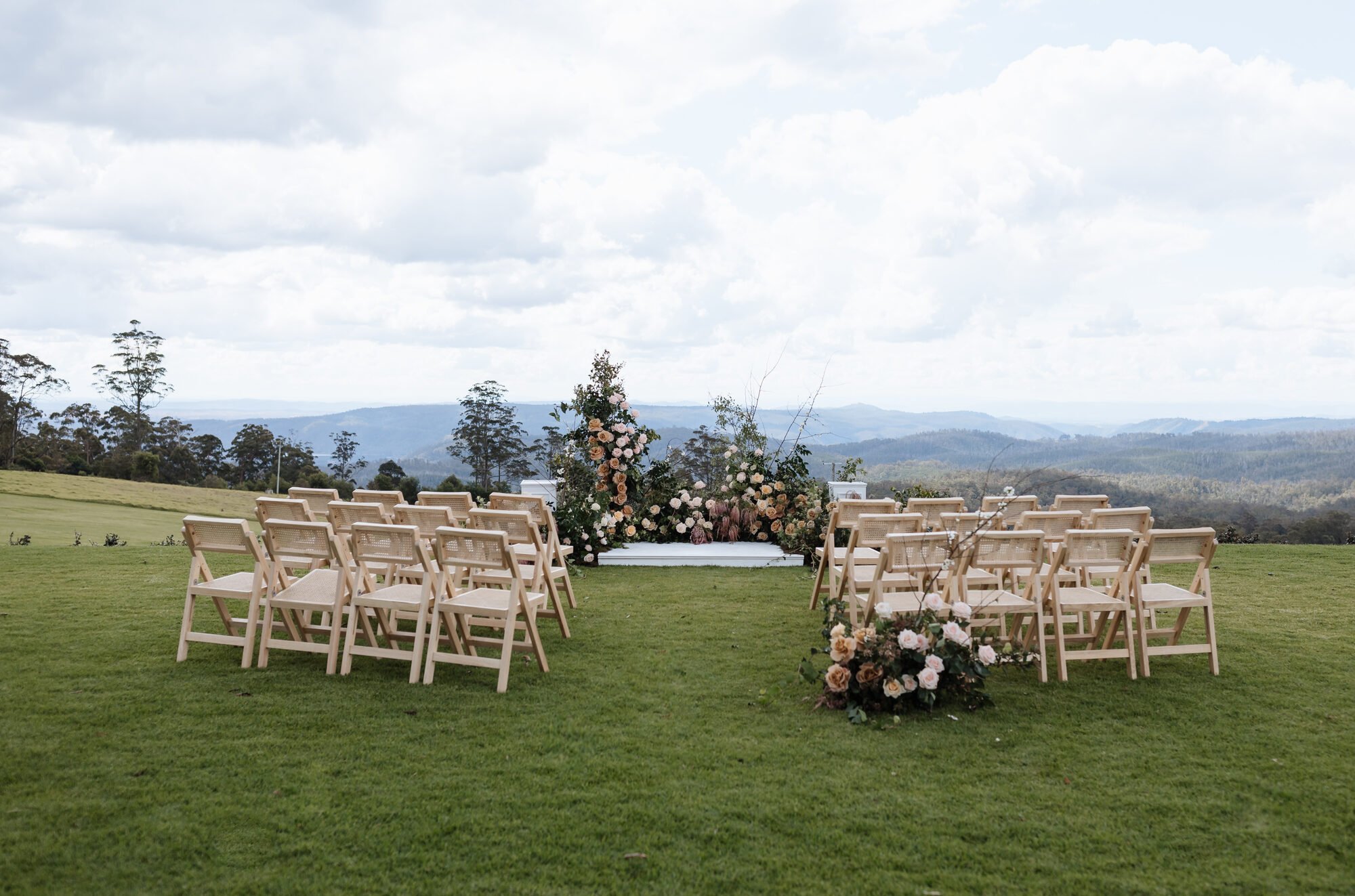 Outdoor wedding ceremony setup on a grassy field with multiple rows of white chairs facing an altar decorated with pink, white, and peach flowers and greenery, overlooking rolling hills and a partly cloudy sky.