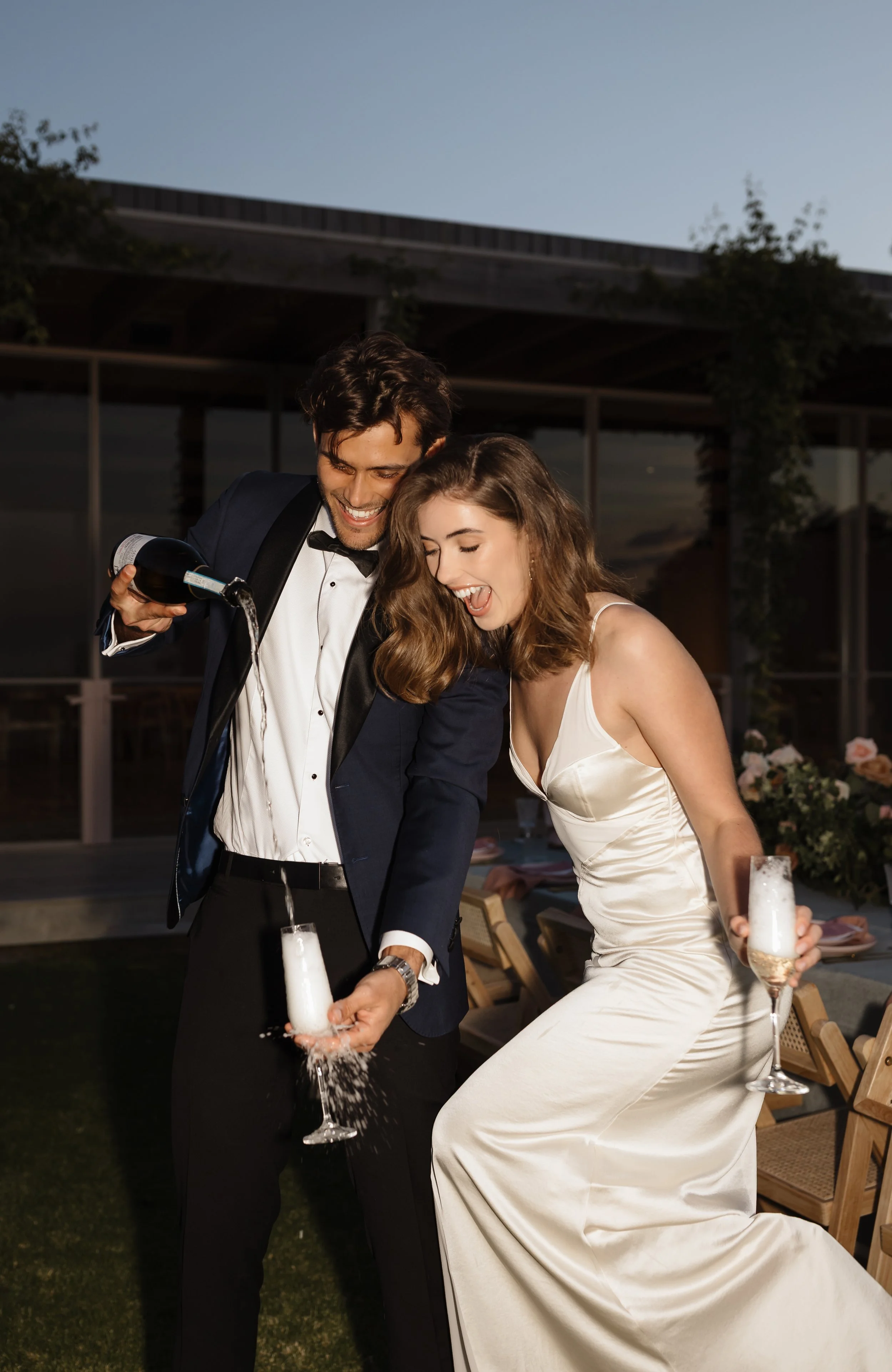 A man and a woman at a celebration, pouring champagne into glasses, with the man in a tuxedo and the woman in a white dress, possibly wedding attire, lit by evening light.