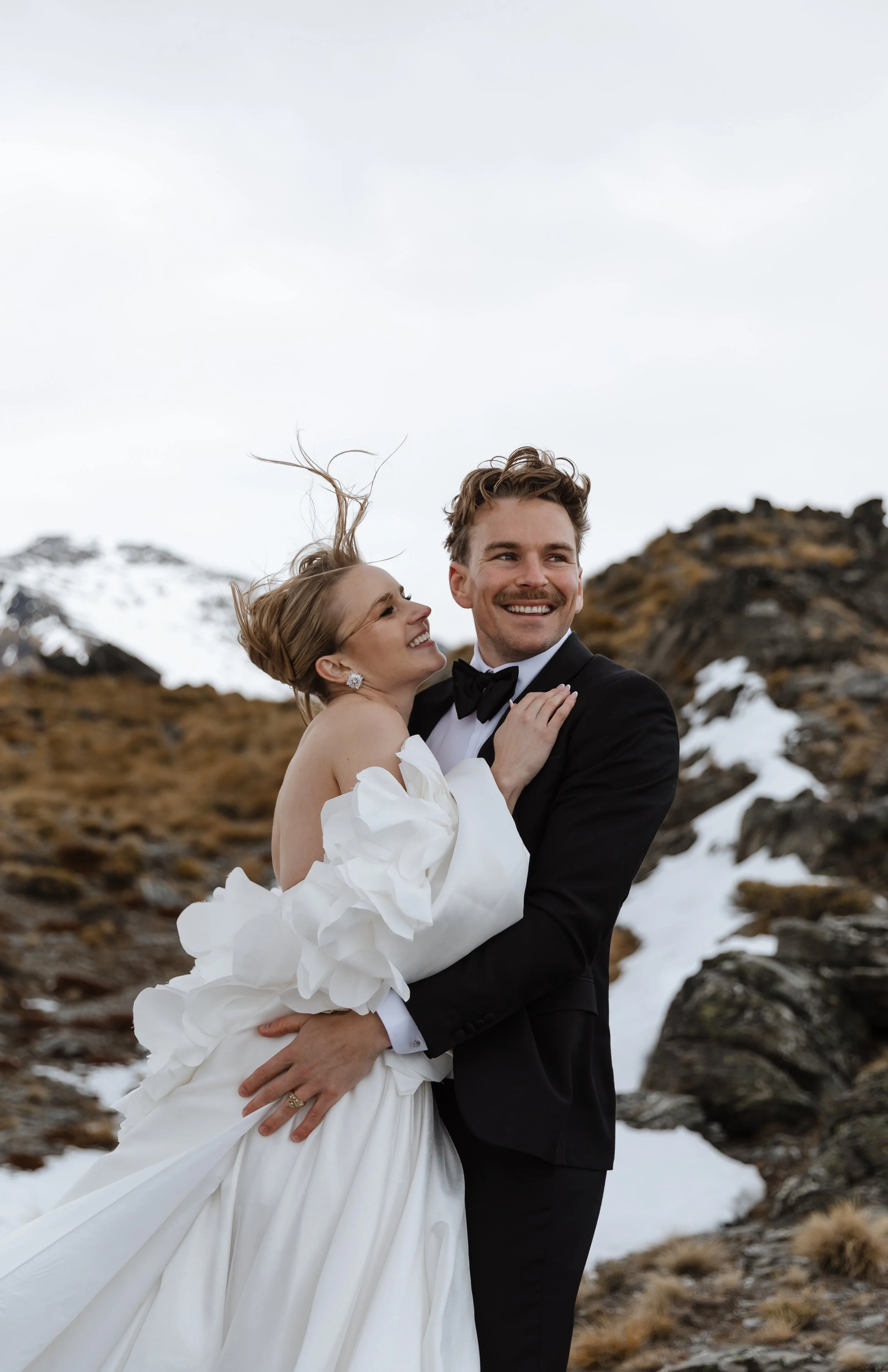 A smiling bride and groom dancing outside on a rocky and snowy mountain terrain, with mountains in the background and overcast sky.