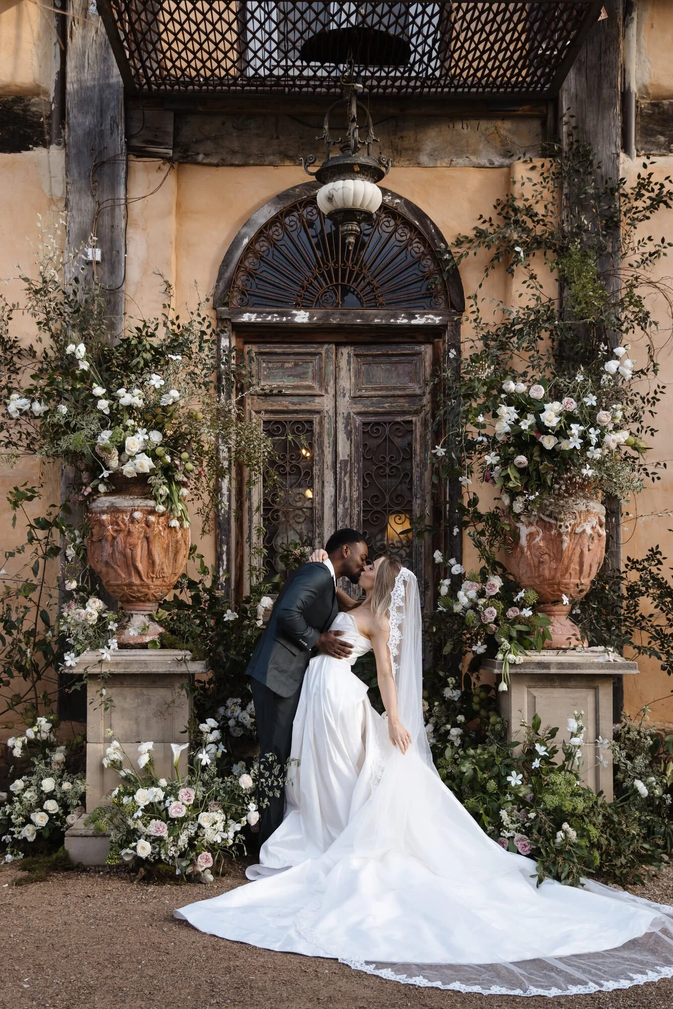 A bride and groom sharing a kiss in front of an ornate, rustic door surrounded by large flower arrangements on columns.