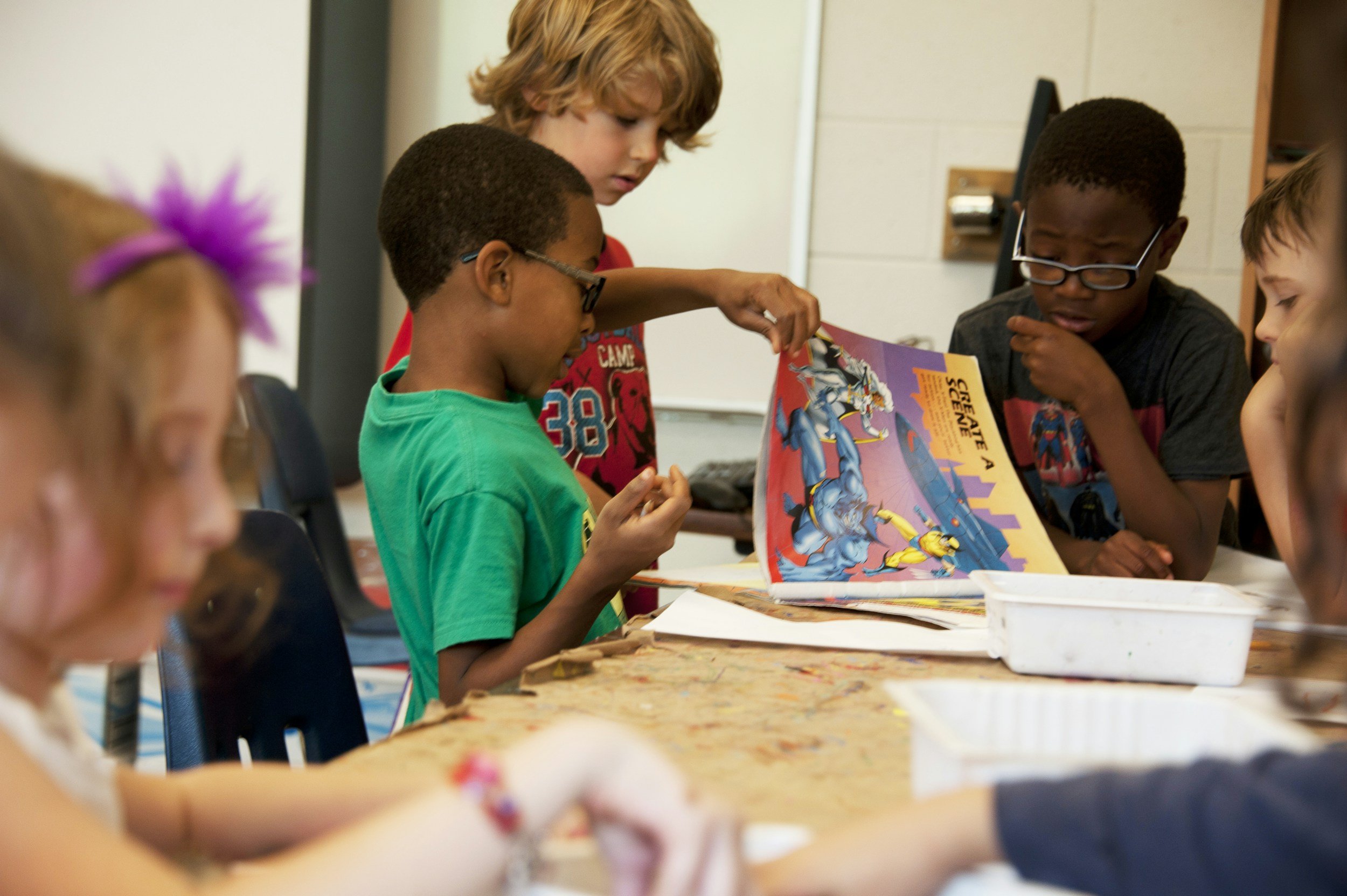Children seated around a table in a classroom looking at a colorful comic book.
