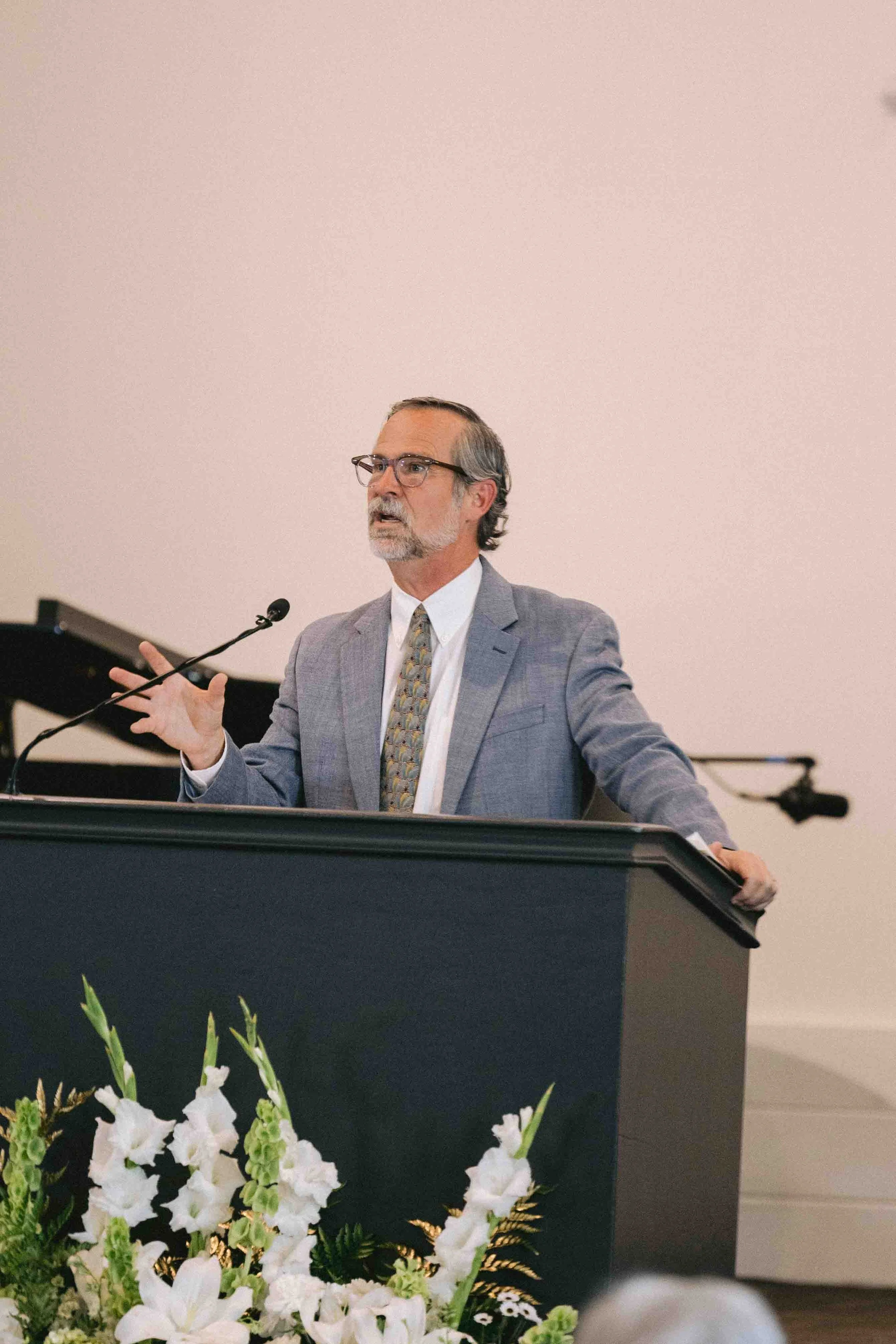 A man wearing glasses and a gray suit stands behind a podium with a microphone, gesturing with his right hand during a presentation. In front of the podium, there is a floral arrangement with white flowers and green leaves. The background is plain and light-colored.