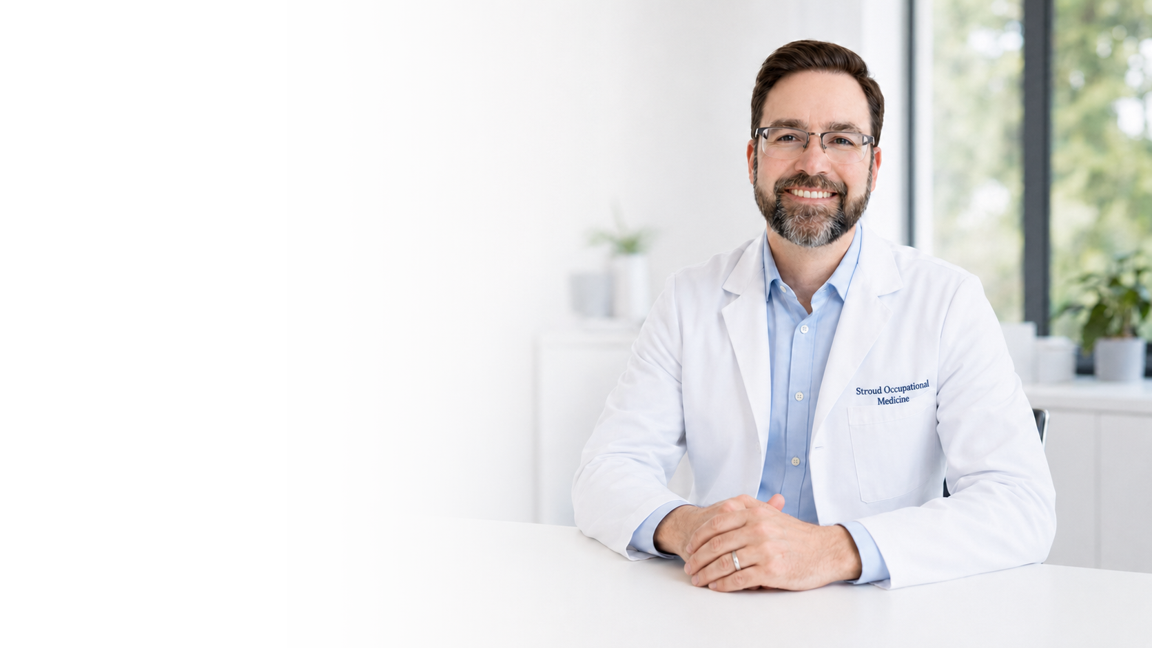 A smiling male doctor with glasses and a beard sitting at a white table in a modern medical office, wearing a white lab coat and a light blue shirt. Large windows in the background let in natural light.