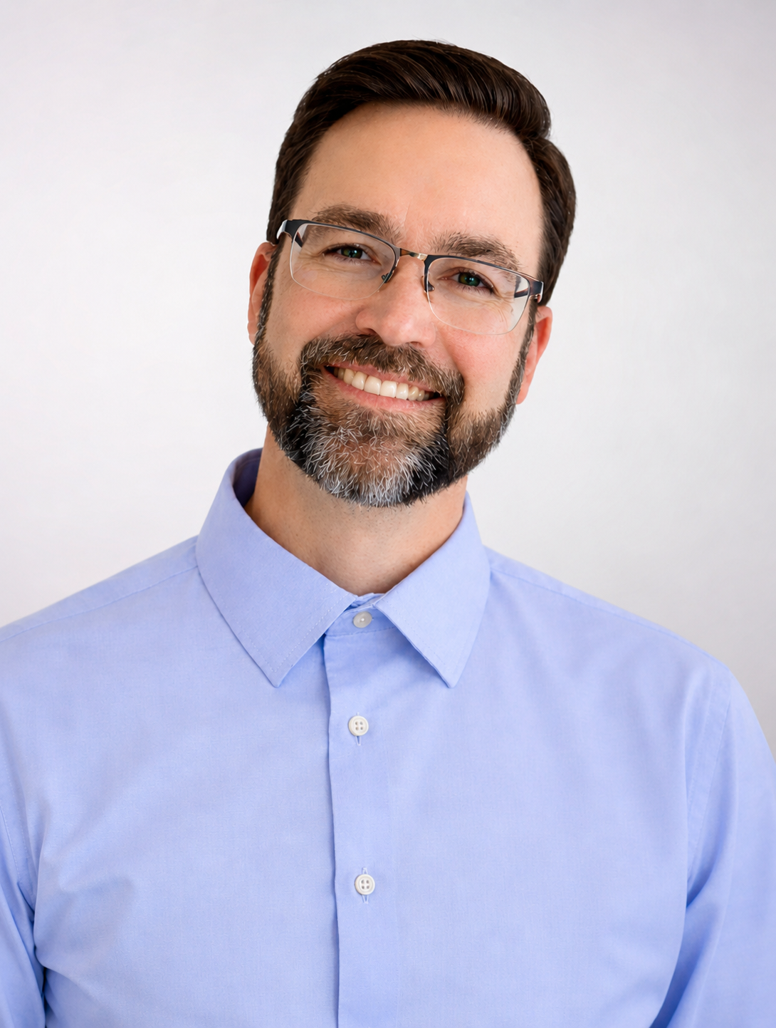 A smiling man with glasses and a beard wearing a light blue dress shirt against a plain background.