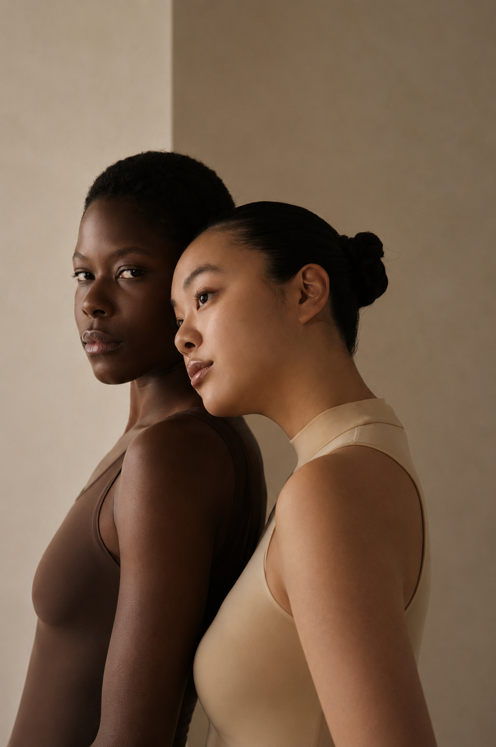 Two women standing close with their heads touching, both wearing sleeveless tops in neutral tones against a plain wall.