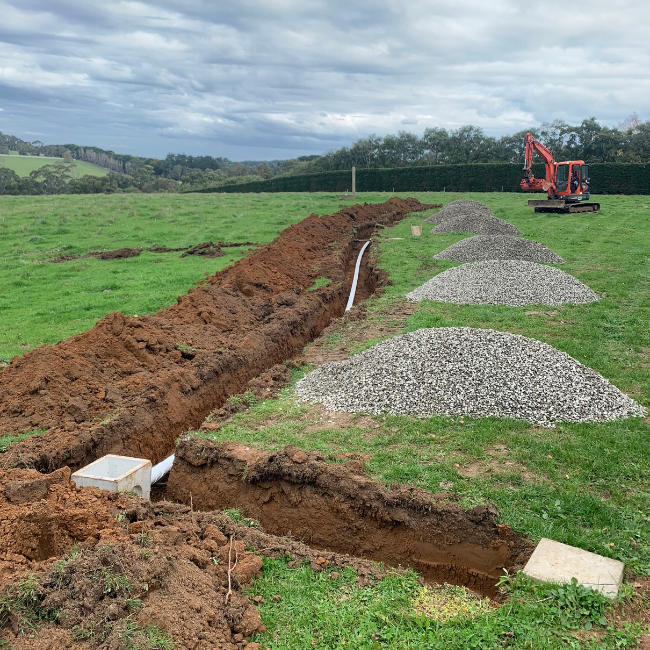 A trench being dug in a grassy field with a small orange excavator nearby. Piles of gravel are stacked along the trench, and a white pipe runs through it. The scene is outdoors with a cloudy sky and a horizon of trees in the background.