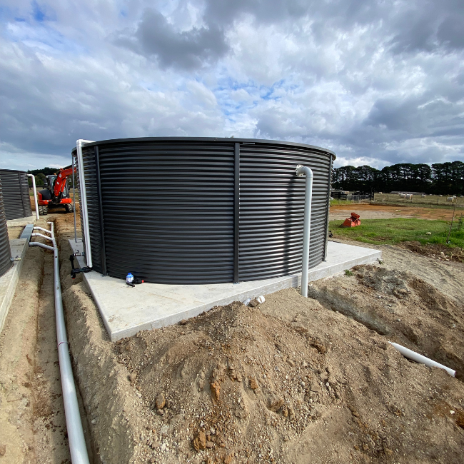 Large black cylindrical water storage tank on a concrete pad at a construction site, with pipes connected to it and partly excavated earth around.