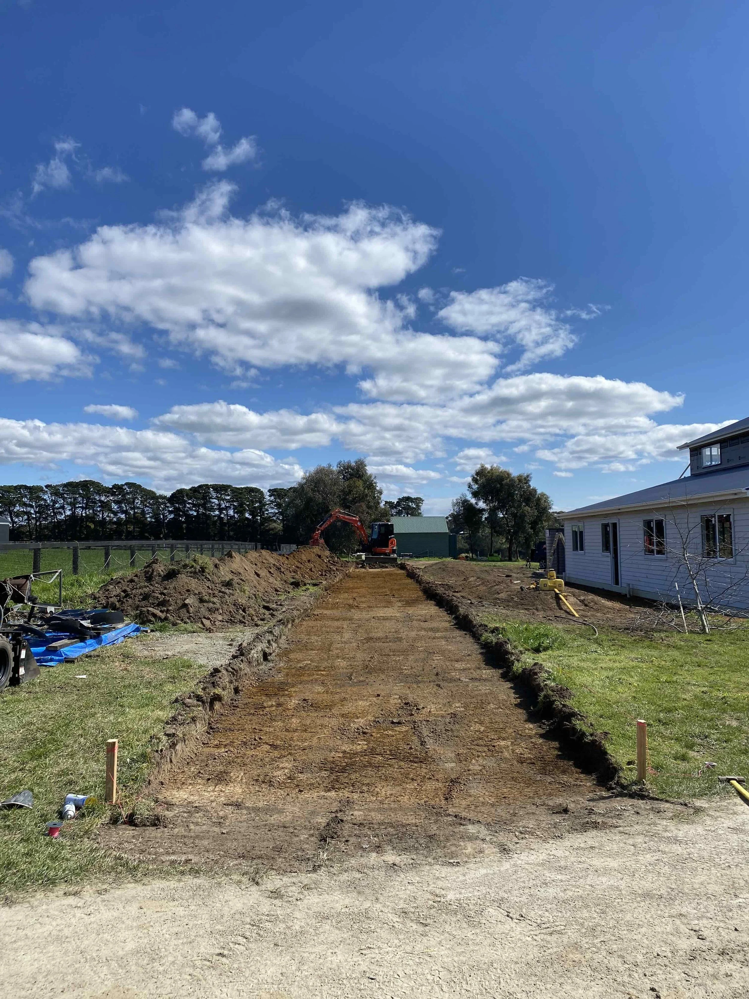 A new driveway being constructed on a rural property. The image shows a straight, level cut ready for gravel to be poured. In the background you see an orange digger.