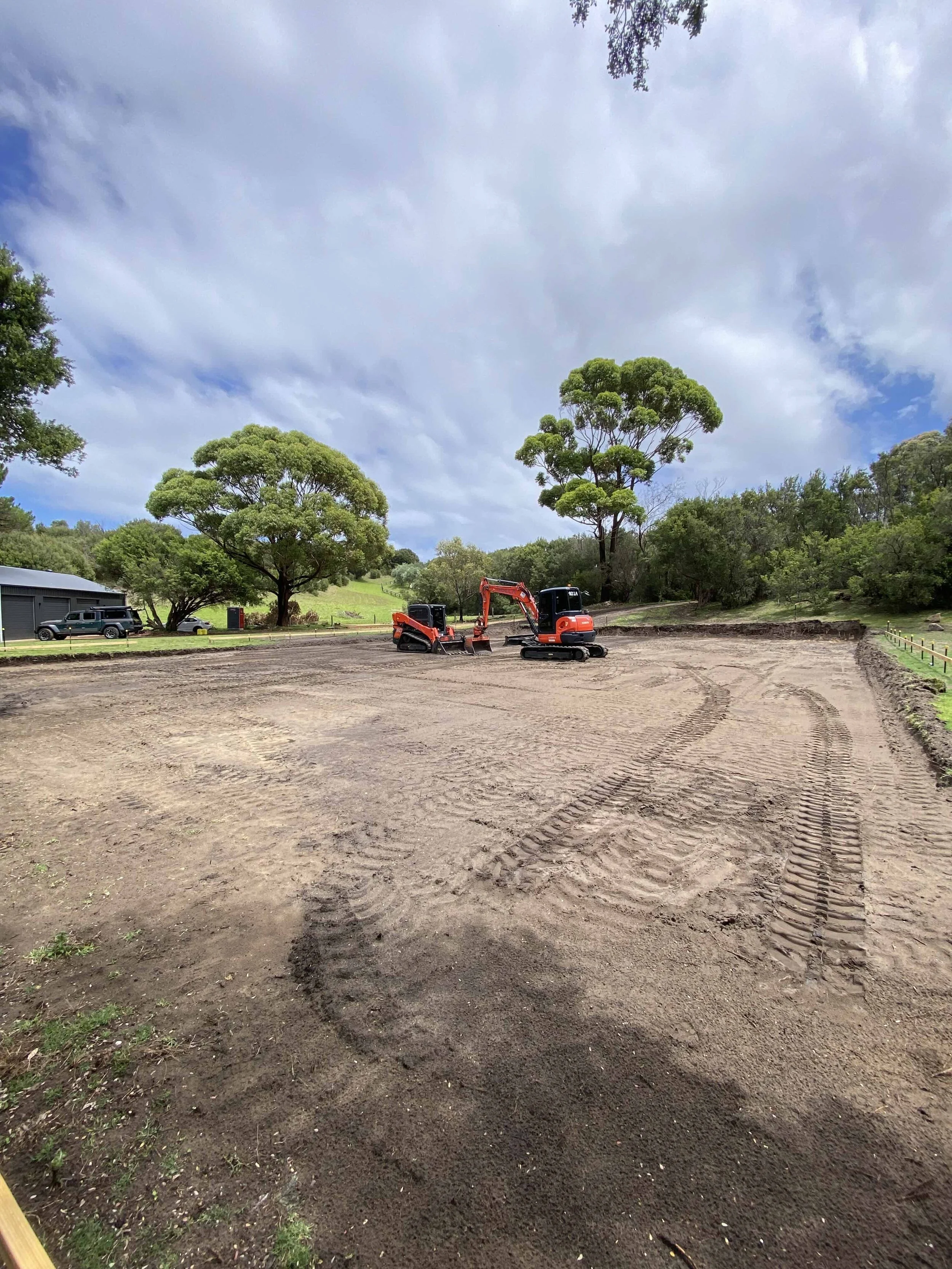 A large site has been prepared. Brown dirt, cut to size and flattened. In the background sits an orange excavator and orange mini bulldozer.