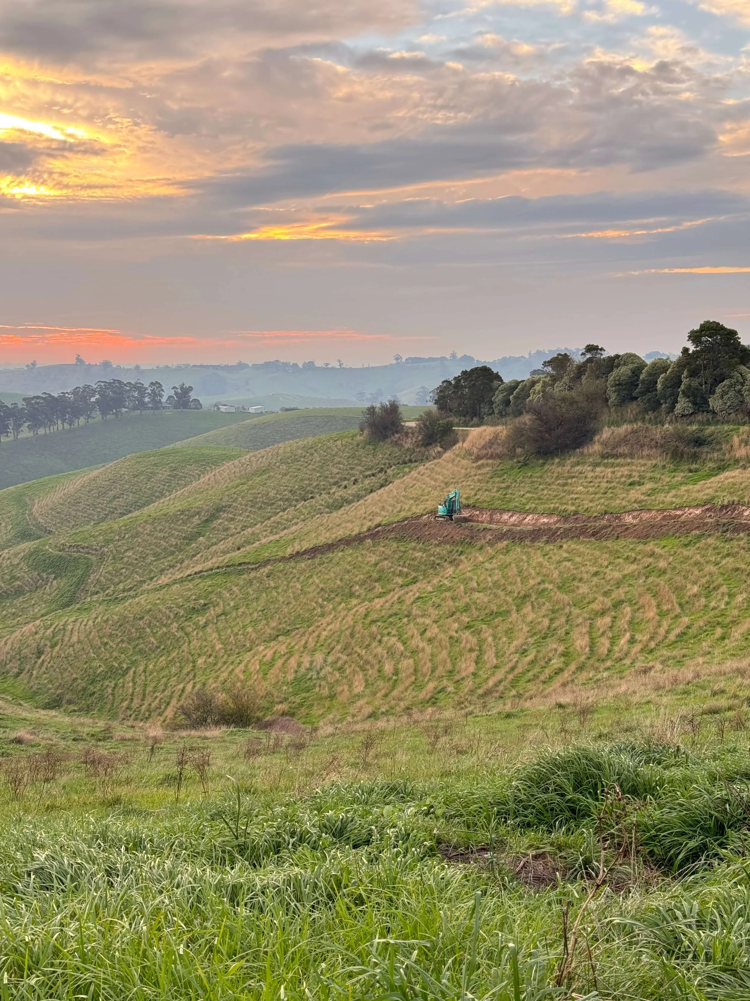 A scenic sunset view from the Strzelecki foothills in Yarragon South. In the distance a blue Rolla Constructions excavator has been working on a farm access road. The green hills and sunset looking into the valley is rural West Gippsland.
