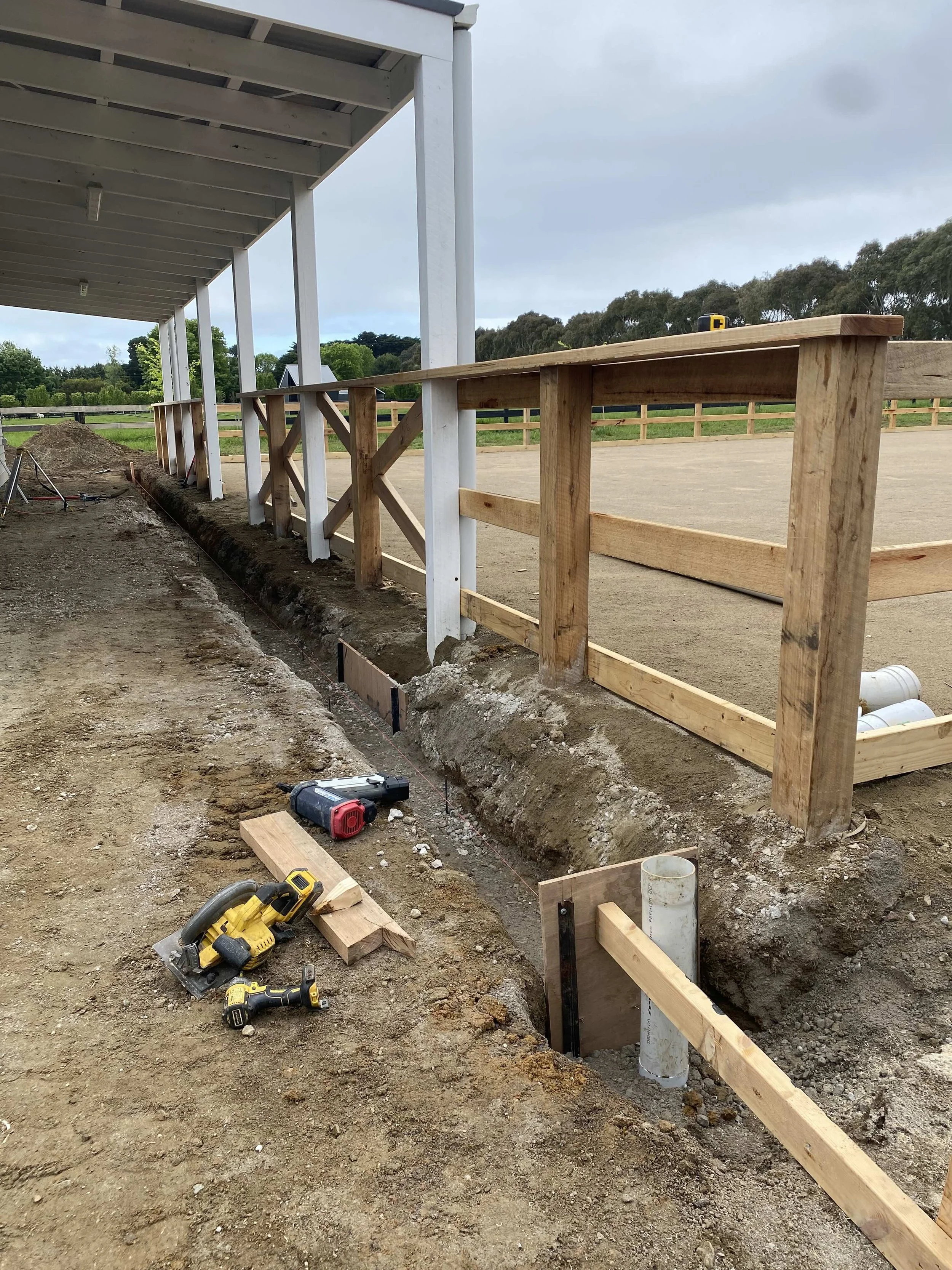 Rolla Constructions installing drainage around a brand new horse arena being built. Tools in foreground of trench. Background wooden fence and horse arena.