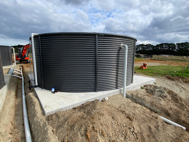 Brand new black water tank installed on a concrete pad on raised ground and surrounded by pipes - indicating a new tank installation.