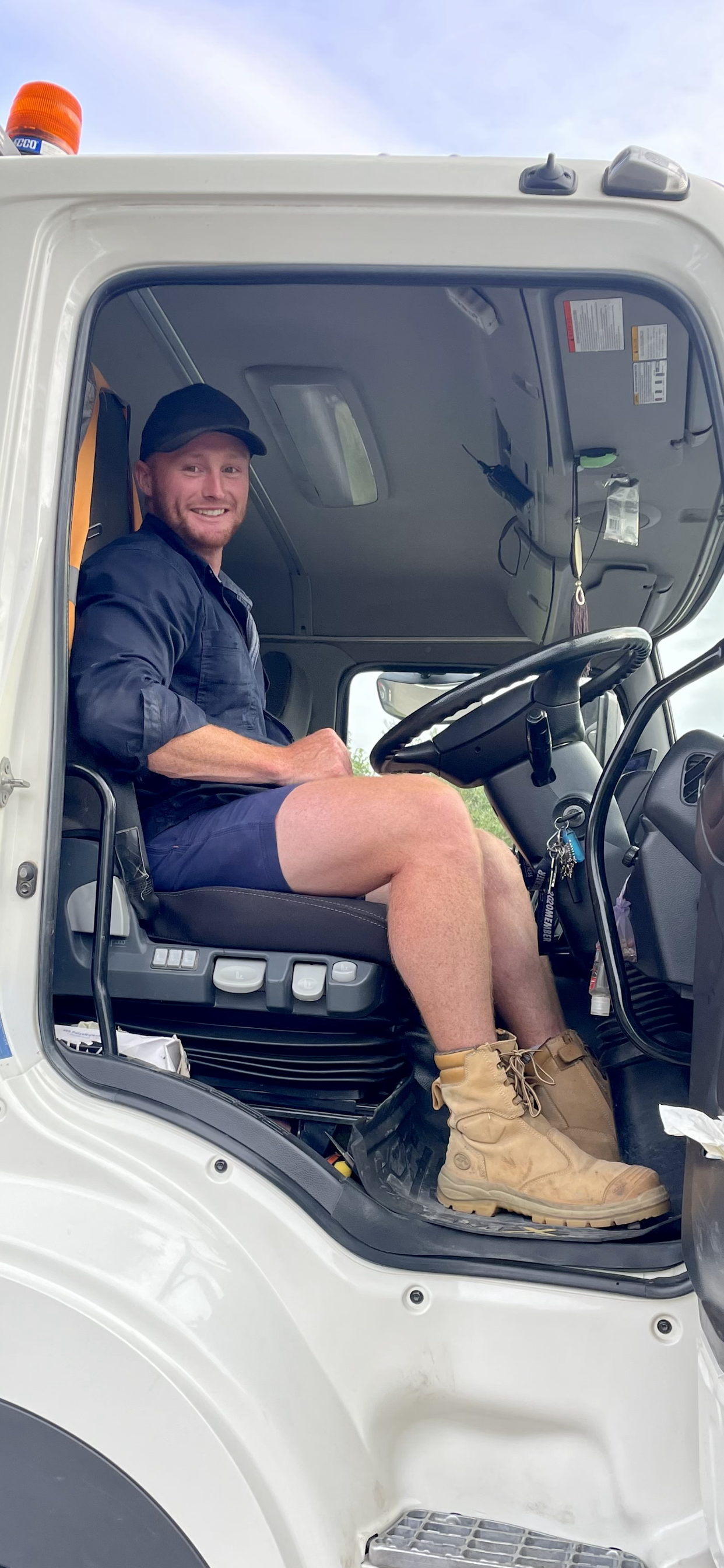 Jake Tayler smiling from the truck cabin. He's in blue workwear shorts and has tan work boots and sits behind the wheel.