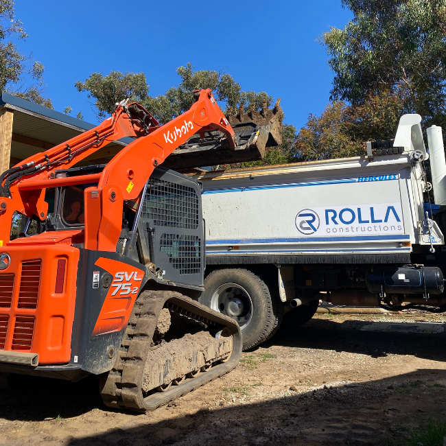 An orange Kubota SVL 75-2 compact track loader lifting a bale of wood or similar material into a white ROLLA construction truck. The scene is outdoors with blue sky and trees in the background.