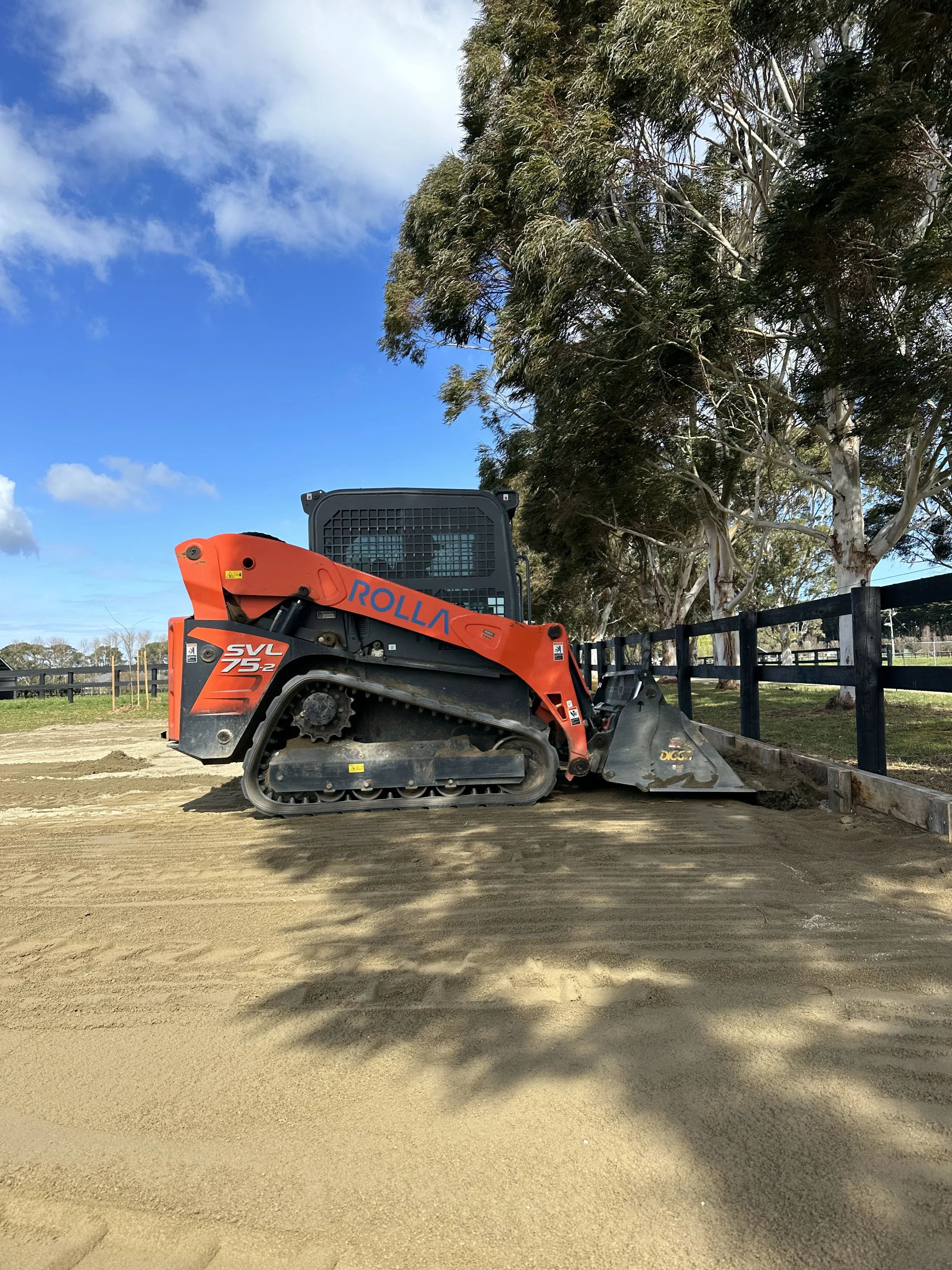 A small orange and black tracked construction vehicle, likely a compact skid steer loader, on a dirt surface next to a black wooden fence, with tall trees and a blue sky with clouds in the background.