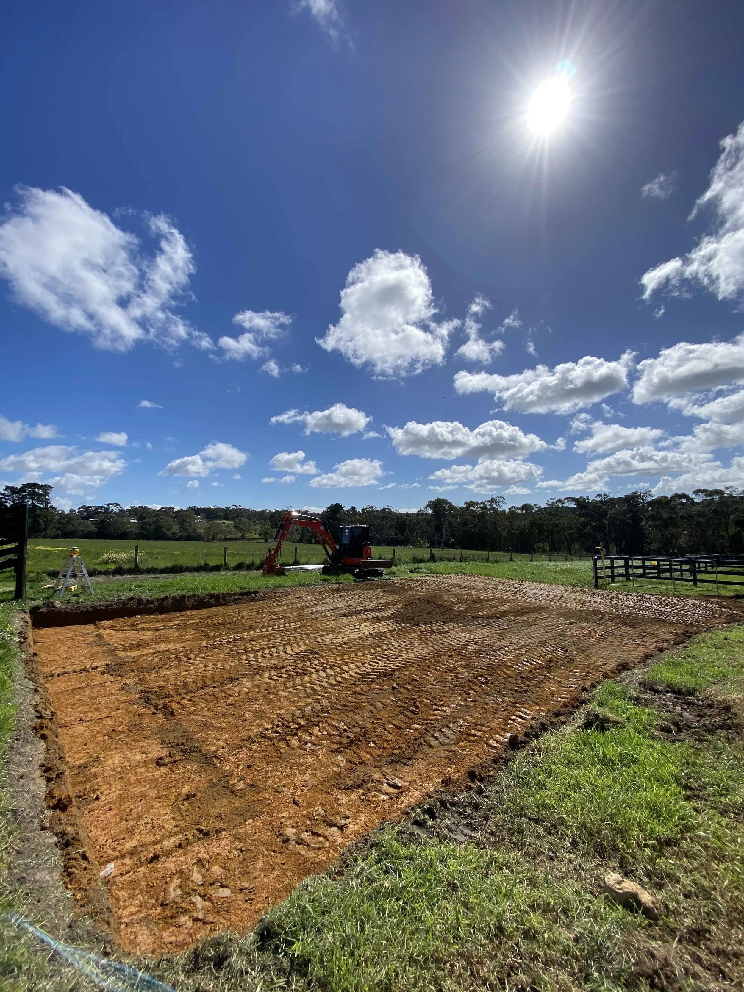 On a rural property or farm surrounded by green paddocks a square site cut with red compacted earth and in the background surveying equipment.