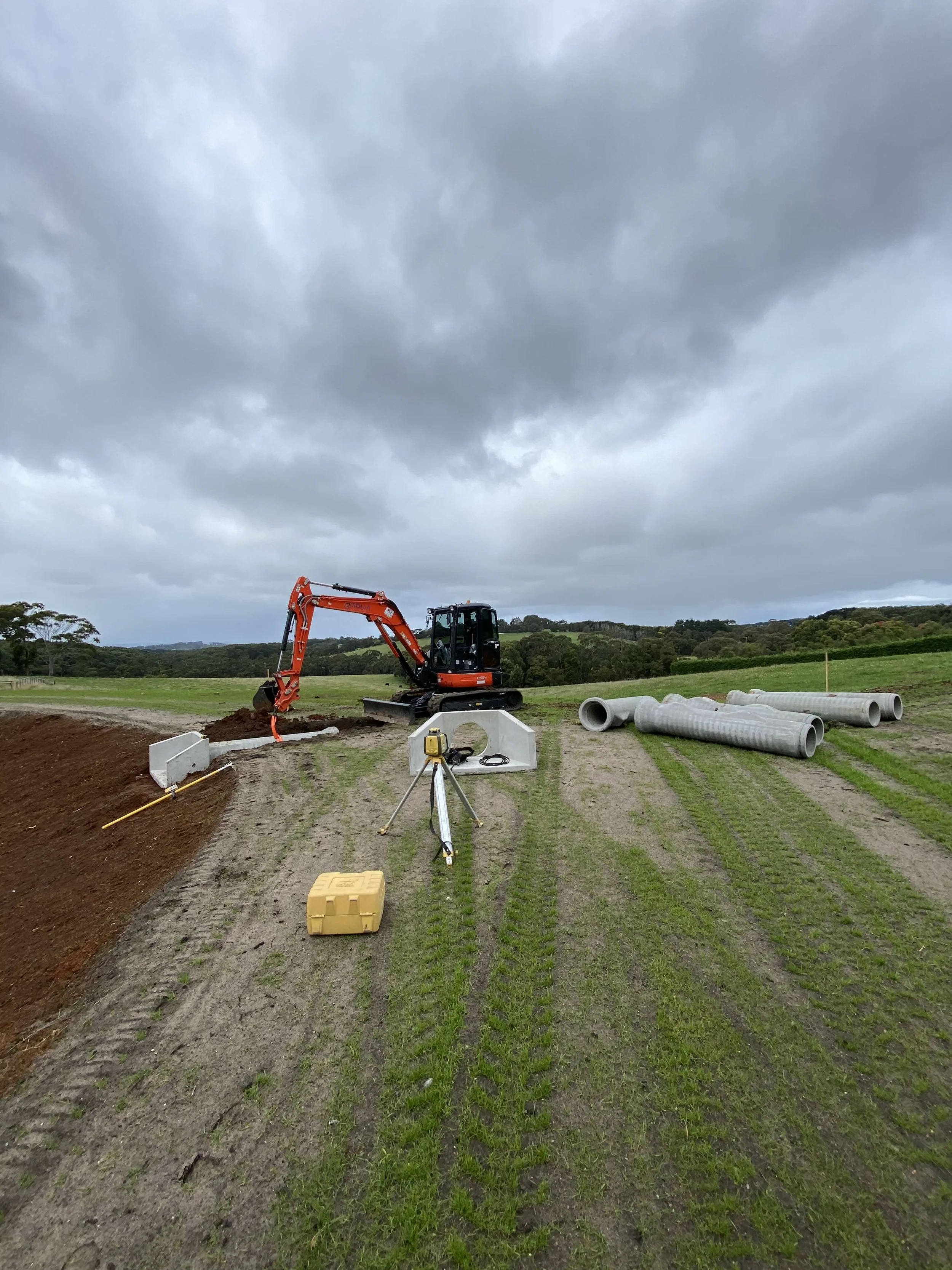Construction site in a grassy field with dug soil, a small orange excavator, pipes, surveying equipment, and cloudy sky.
