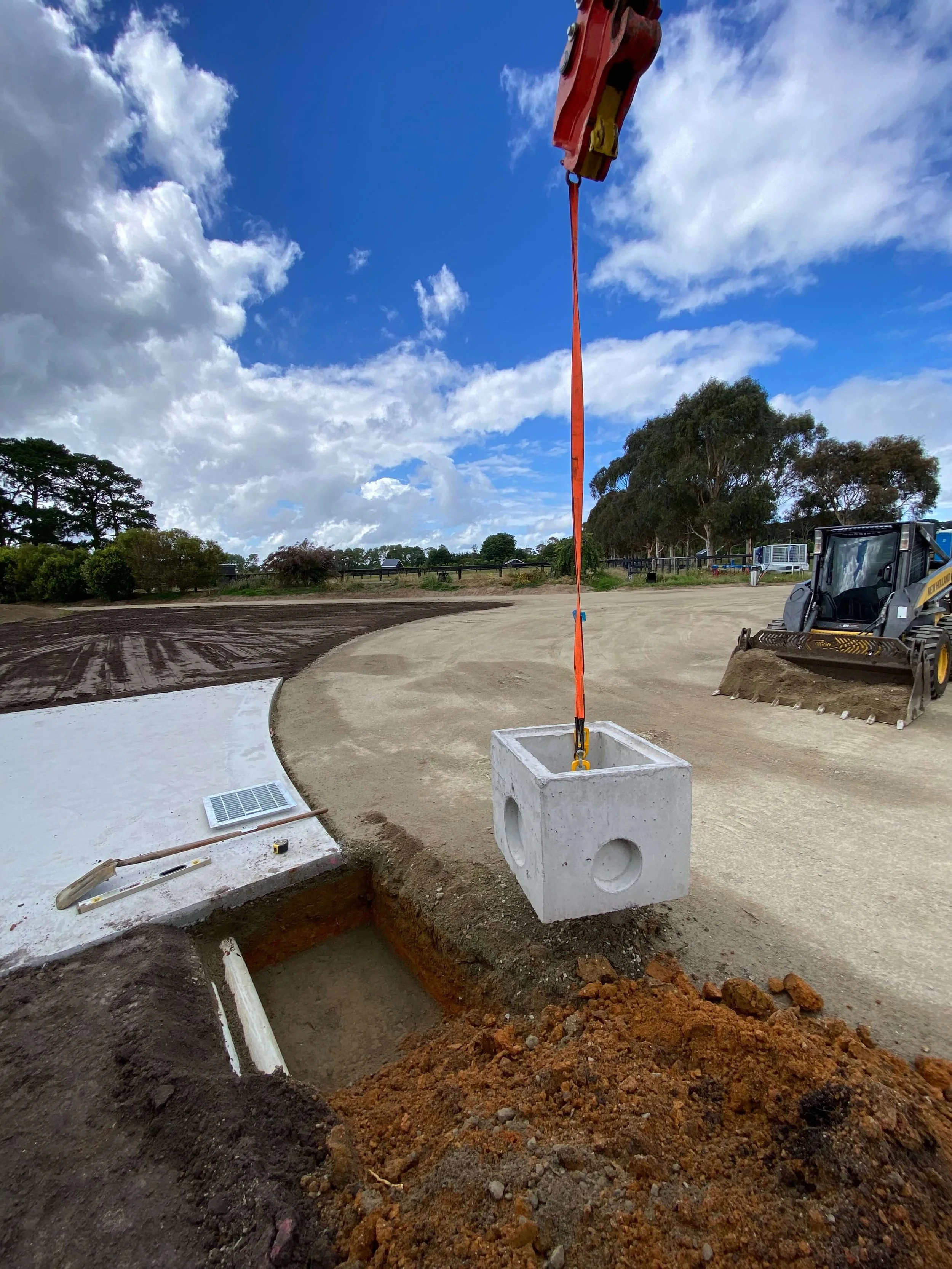 Construction area at a rural lifestyle property. Drainage is being installed alongside the new driveway. In the foreground a concrete drain is being dropped in by an overhead excavator. The site is neatly prepared. A small bulldozer present.