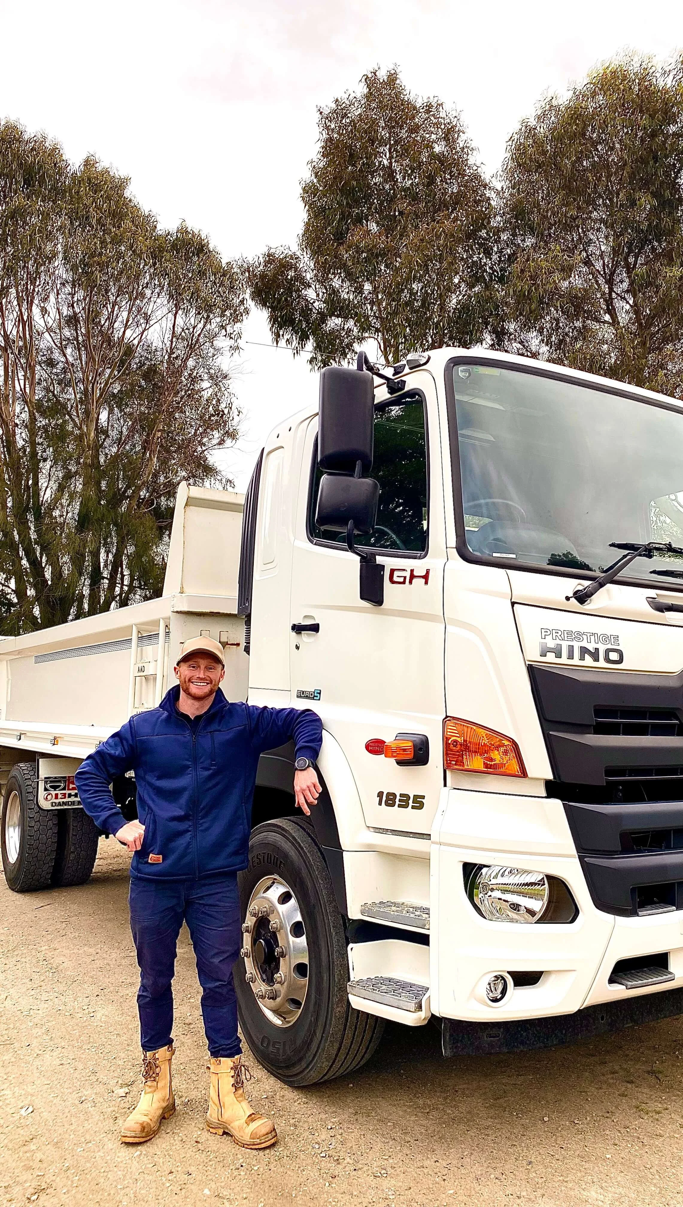 Jake Tayler smiling in a blue jacket, blue pants, and tan boots standing next to a large white Hino truck on a gravel surface with trees in the background.