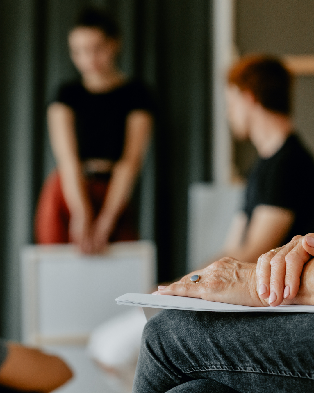Person's hands holding papers in a support group meeting, with a blurred woman standing in the background.