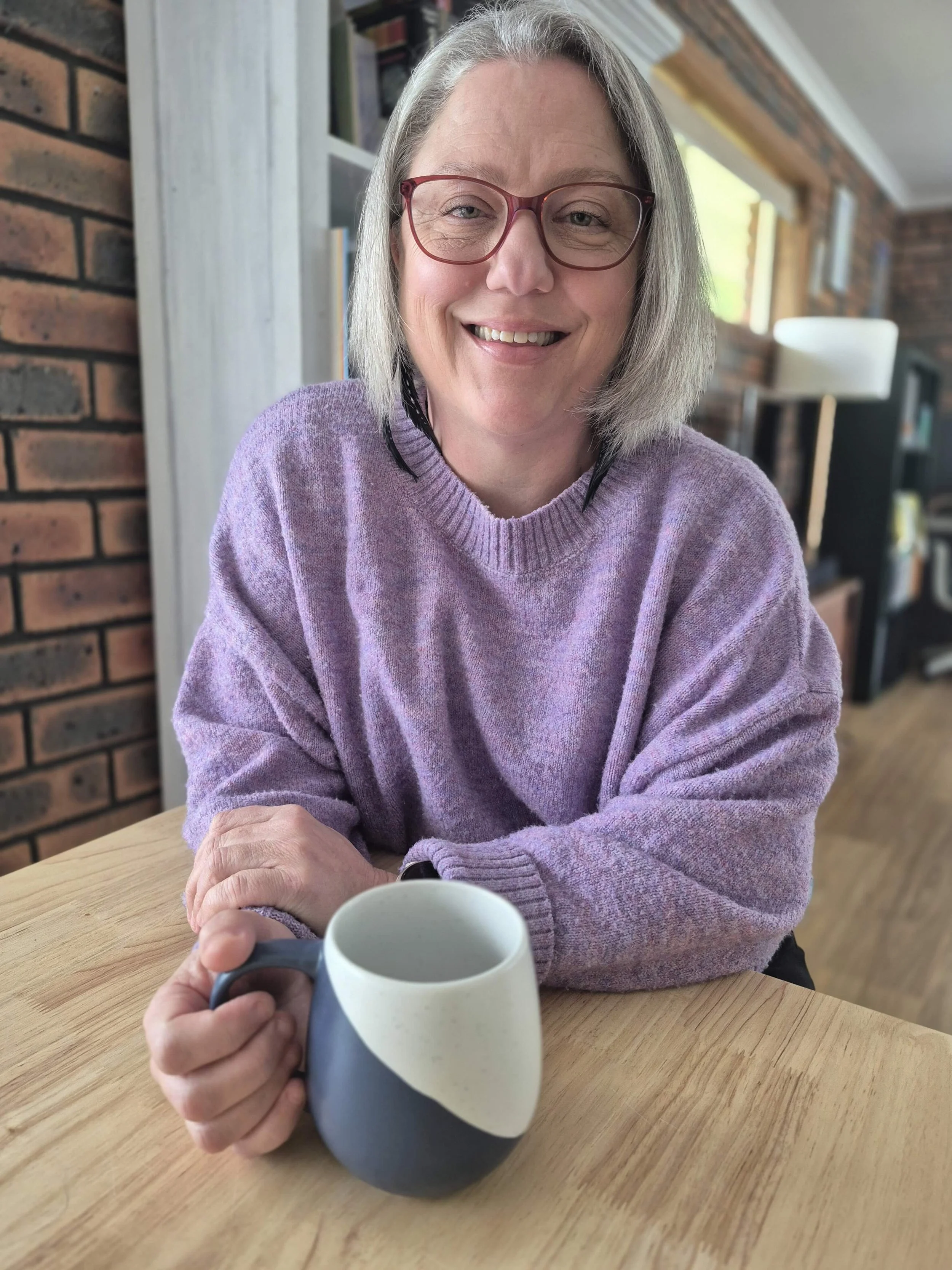 Renée Menzies smiling at camera. She sits at a table, is wearing a purple jumper, has grey hair in a bob and wears maroon glasses. She's holding a mug.