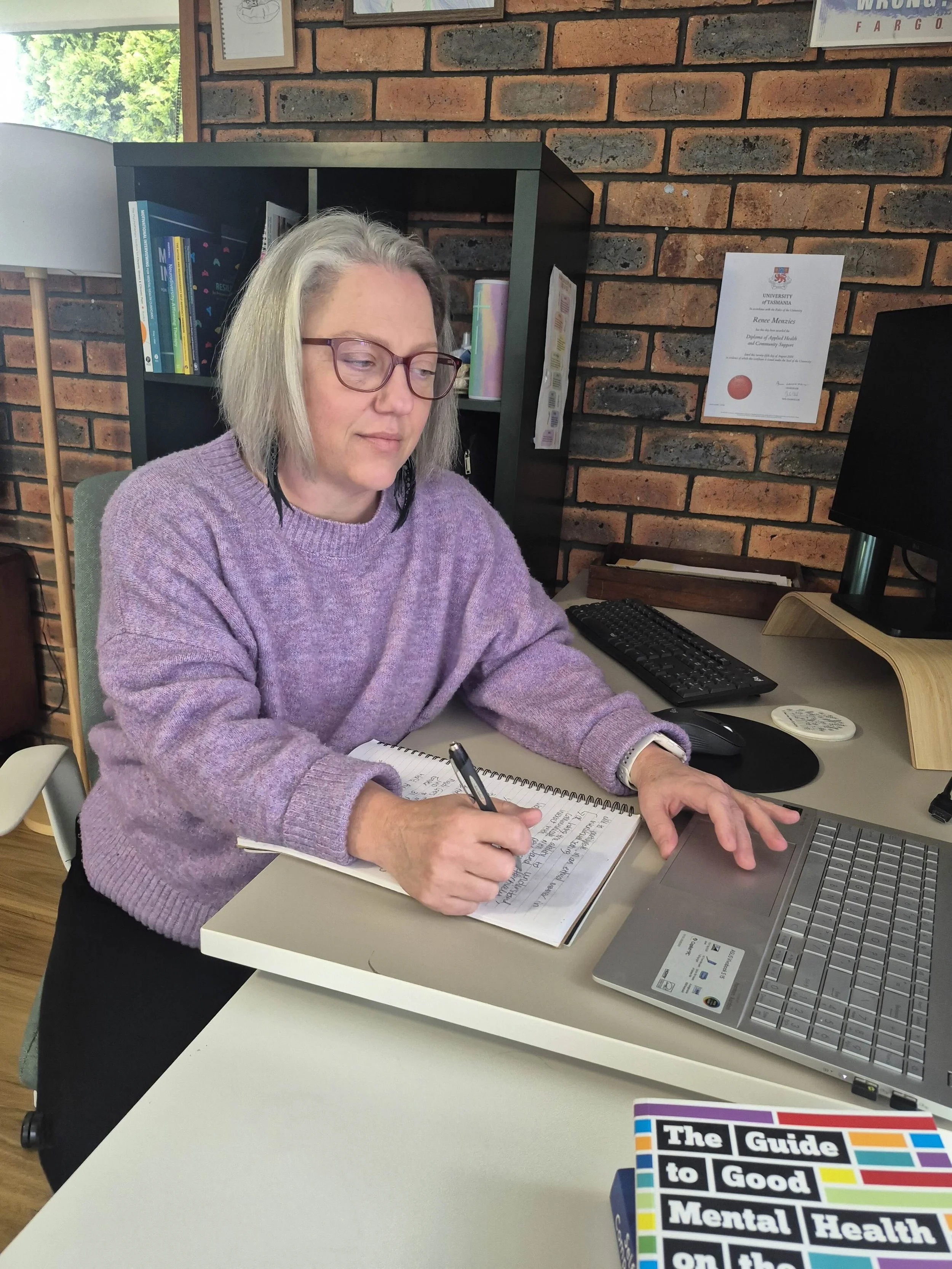 Renée Menzies sits at her desk writing in a notebook and looking at her silver laptop. She's wearing a purple jumper and has grey hair cut to a bob and wears red glasses. In the back ground we see a black bookshelf, exposed brick walls and a Diploma.