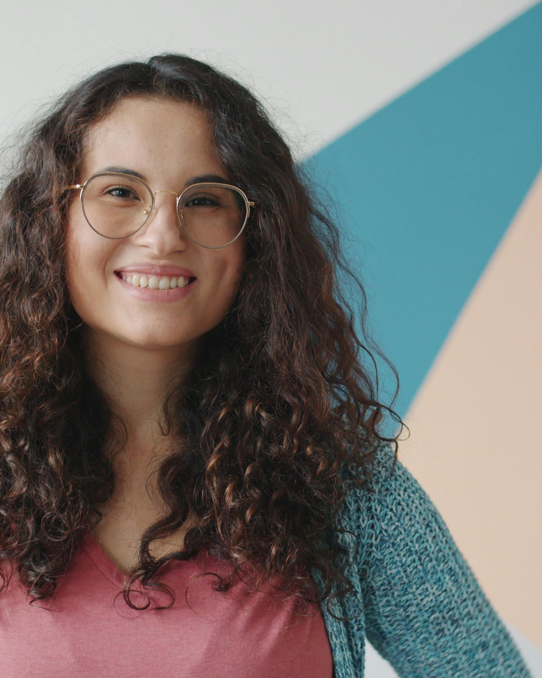 A young woman with long curly hair wearing glasses, a pink shirt, and a blue cardigan, smiling in front of a colorful geometric background.