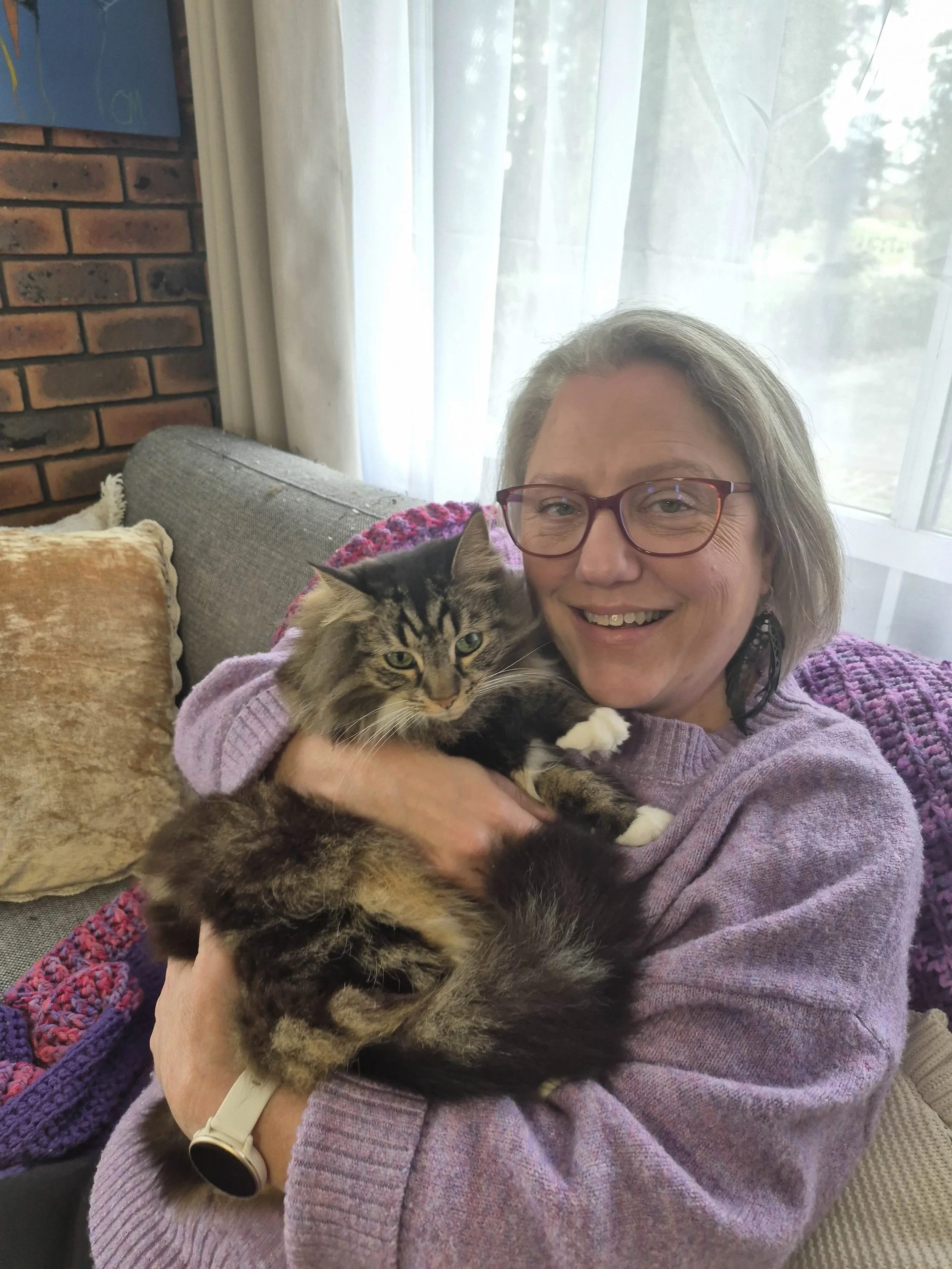Renée Menzies sits on her couch wearing a purple jumper and red glasses. Se has grey hair in a bob and is smiling. She is hugging her cat Luna who is a long-haired tabby.
