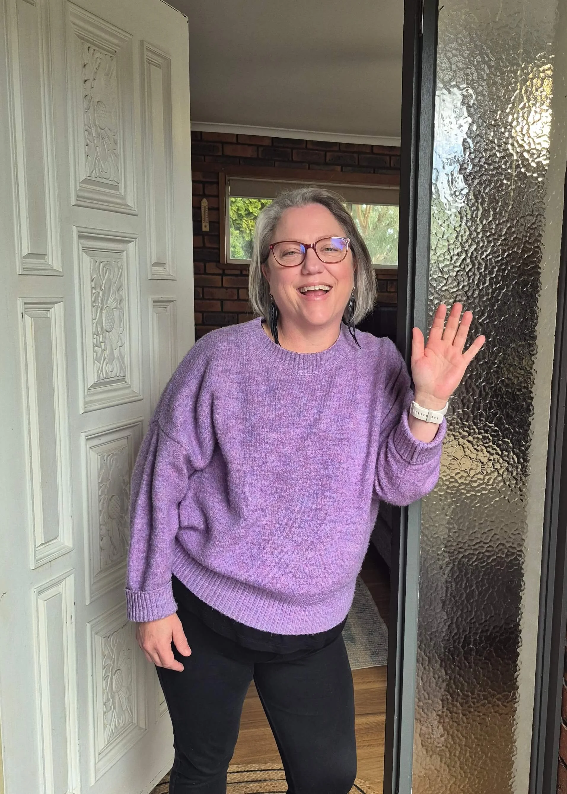 Renée Menzies stands at her front door smiling and waving wearing a purple jumper and red glasses. The door is white with detailed patterns and there is a glass panel on the opposite side. Through the door we see walls with exposed brickwork.