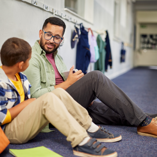 An adult man and a young boy sitting on the floor in a school hallway, engaging in conversation, with lockers and hanging clothing in the background.
