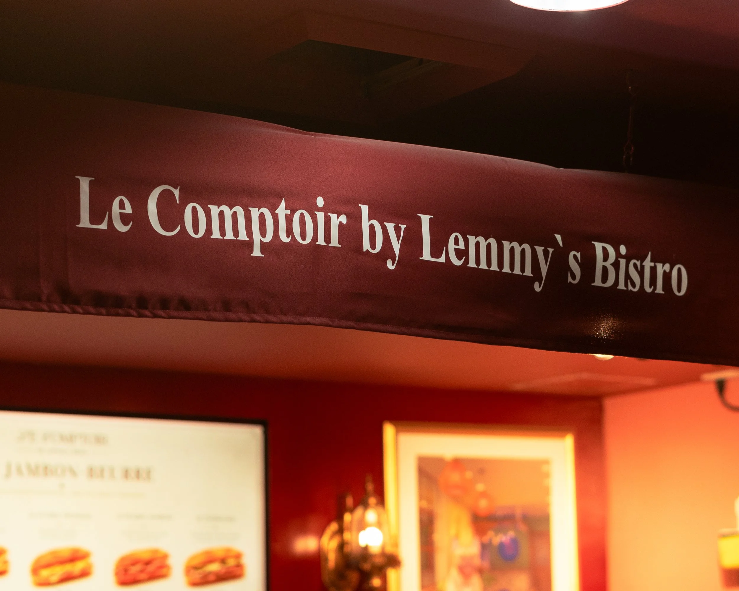 Red sign with white text that reads 'Le Comptoir by Lemmy's Bistro' in a dimly lit restaurant or bar setting.