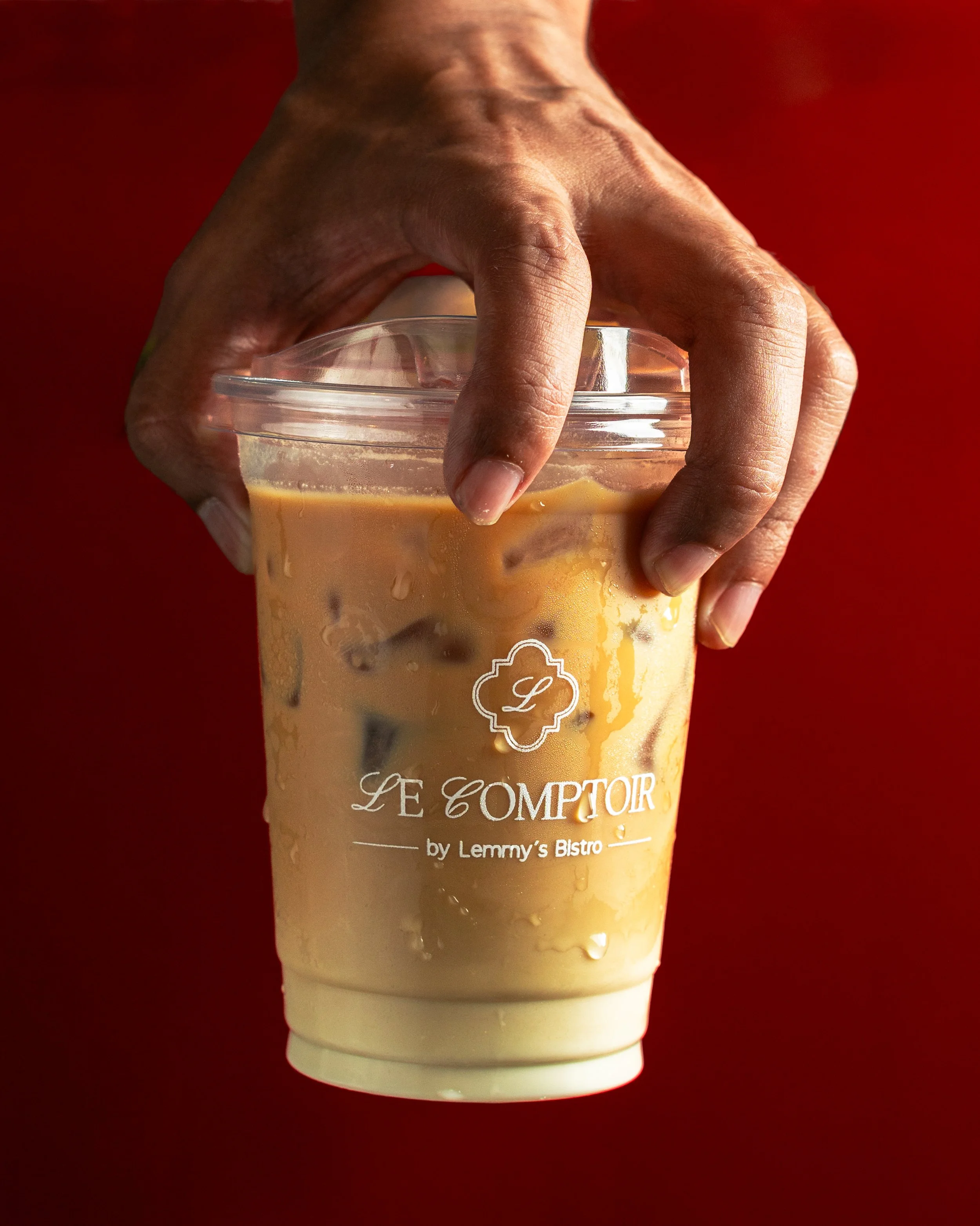 A hand holding a plastic cup filled with iced coffee or latte against a red background. The cup has a lid and features a label that reads 'Le Comptoir by Lemmy's Bistro.'