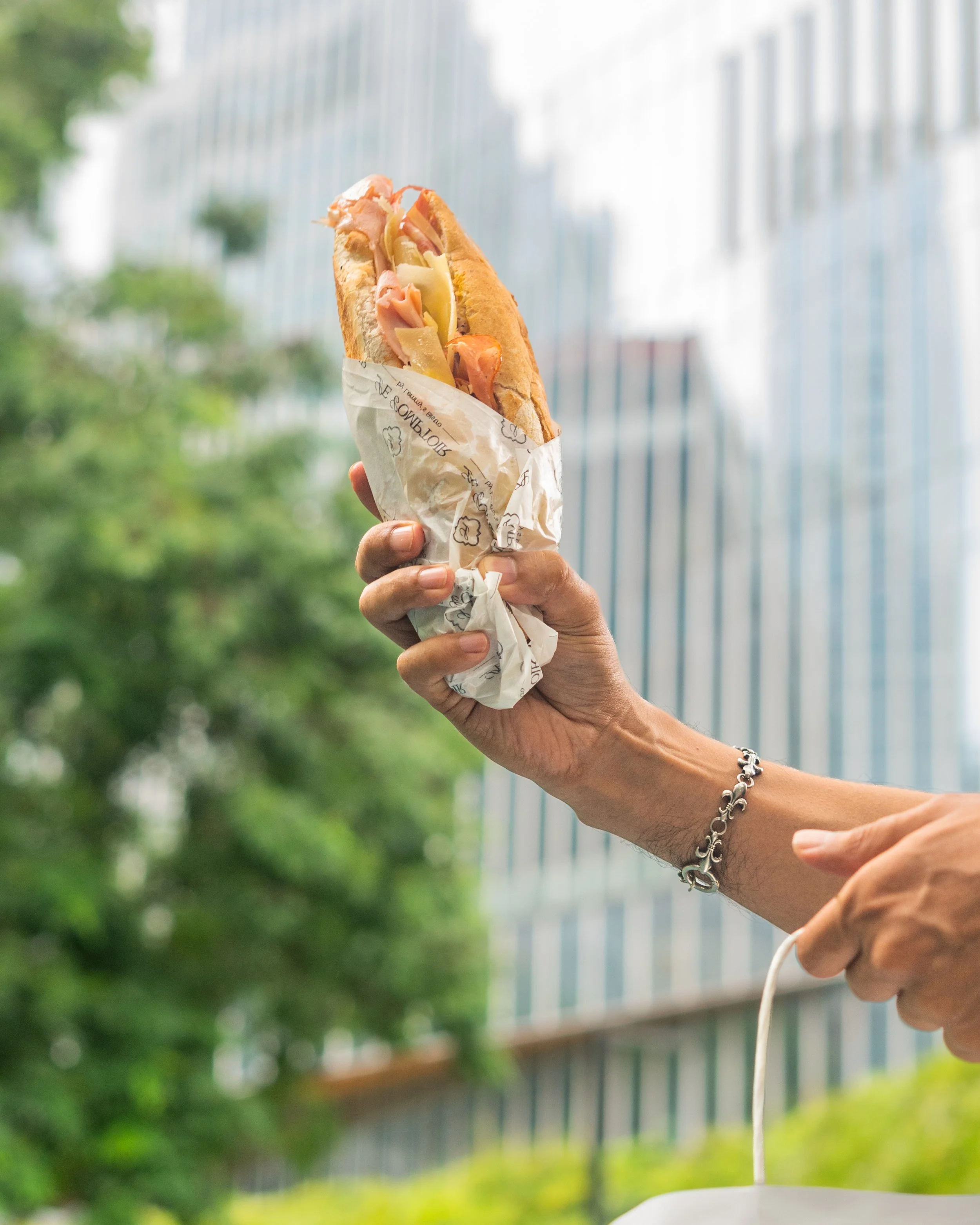 Close-up of a person holding a baguette sandwich filled with ham, cheese, and vegetables, wrapped in branded paper, against an outdoor background with trees and a modern building.
