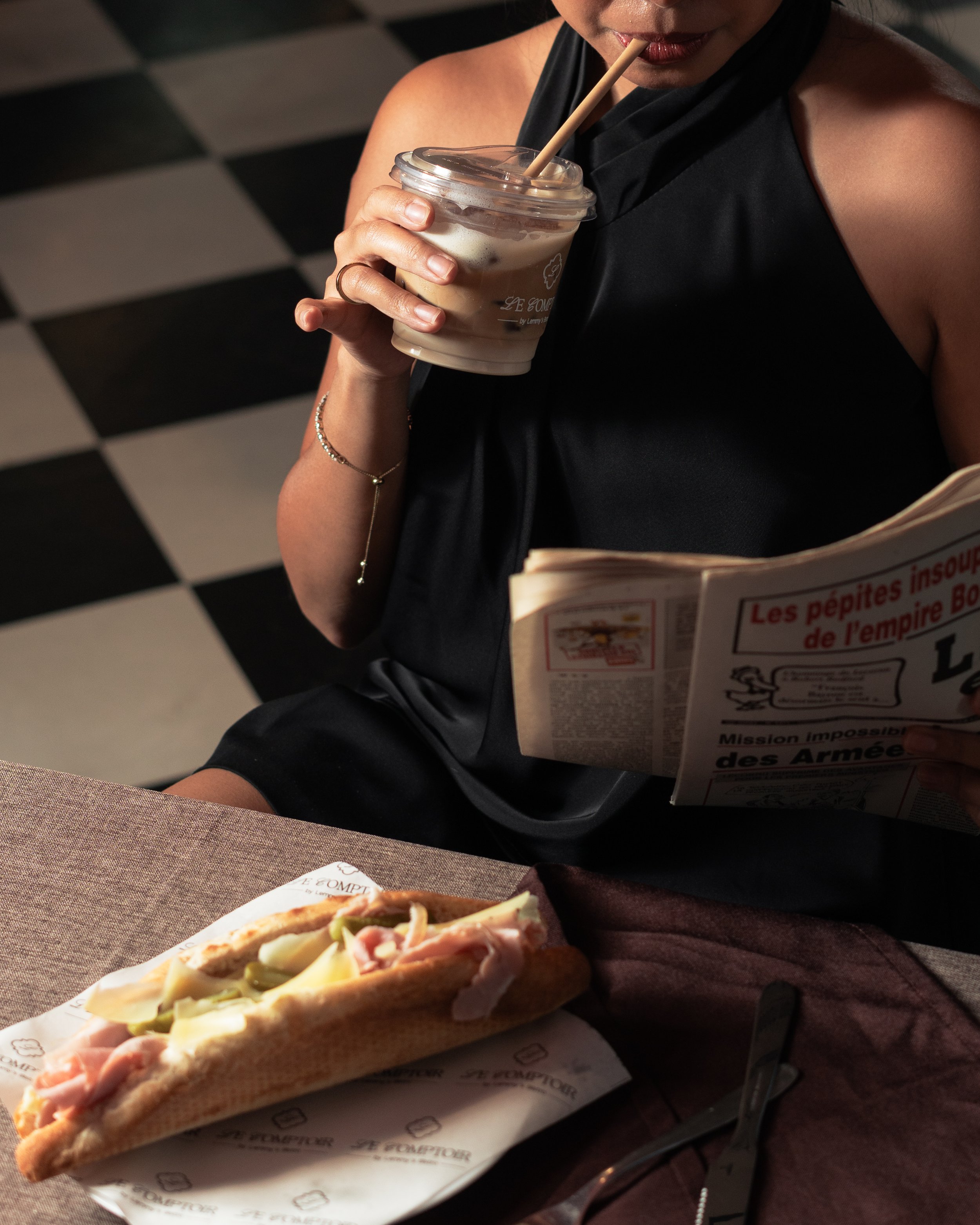 A woman dressed in black holding a large iced coffee with a straw, reading a newspaper, with a sandwich on the table in front of her, in a casual dining setting with checkered floor.