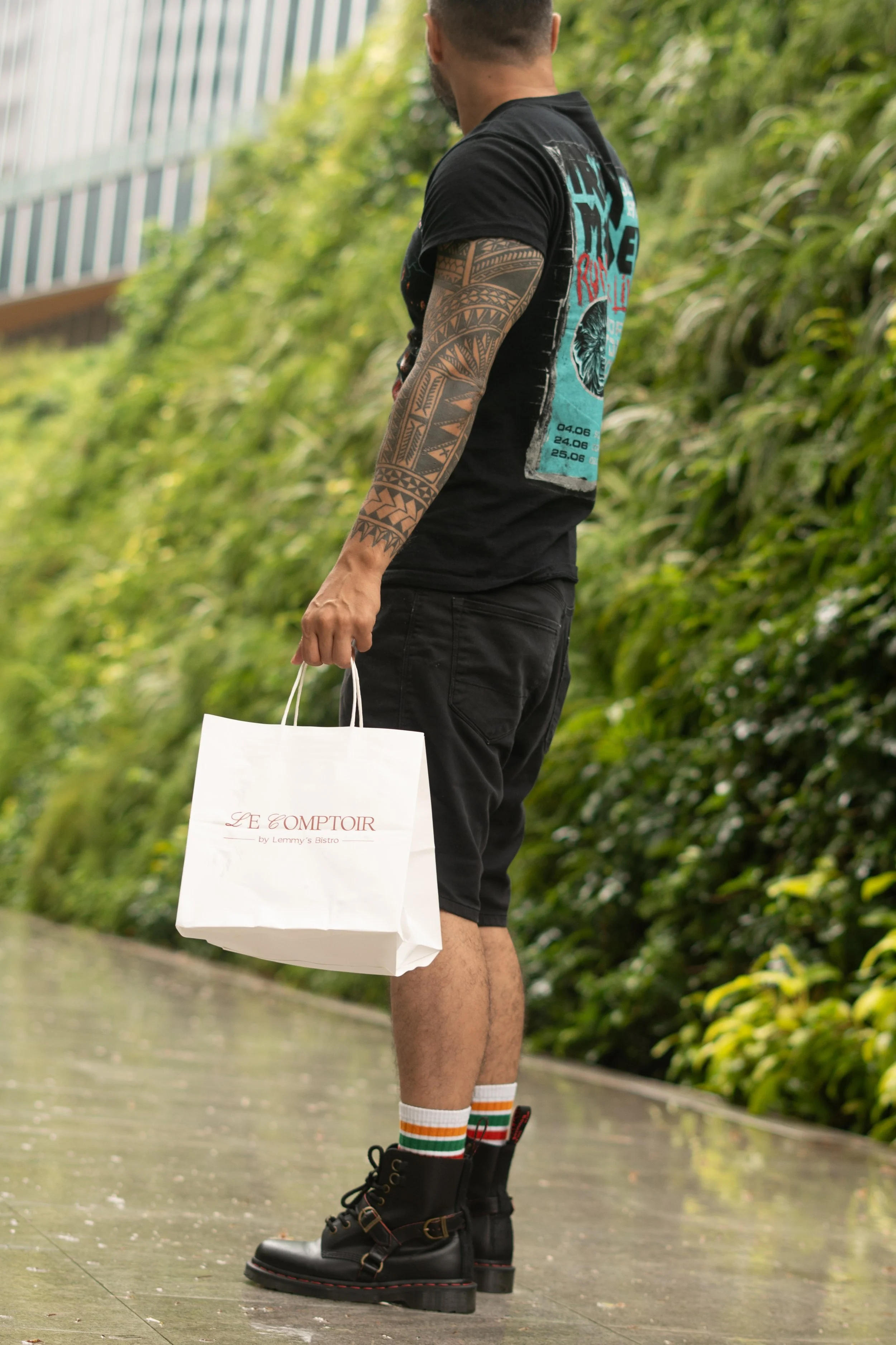 A man standing outdoors on a paved surface holding a white shopping bag with the words 'Le Comptoir' printed on it. He is wearing black shorts, black boots, striped socks, and has a tattooed sleeve on his left arm. The background includes lush greenery.