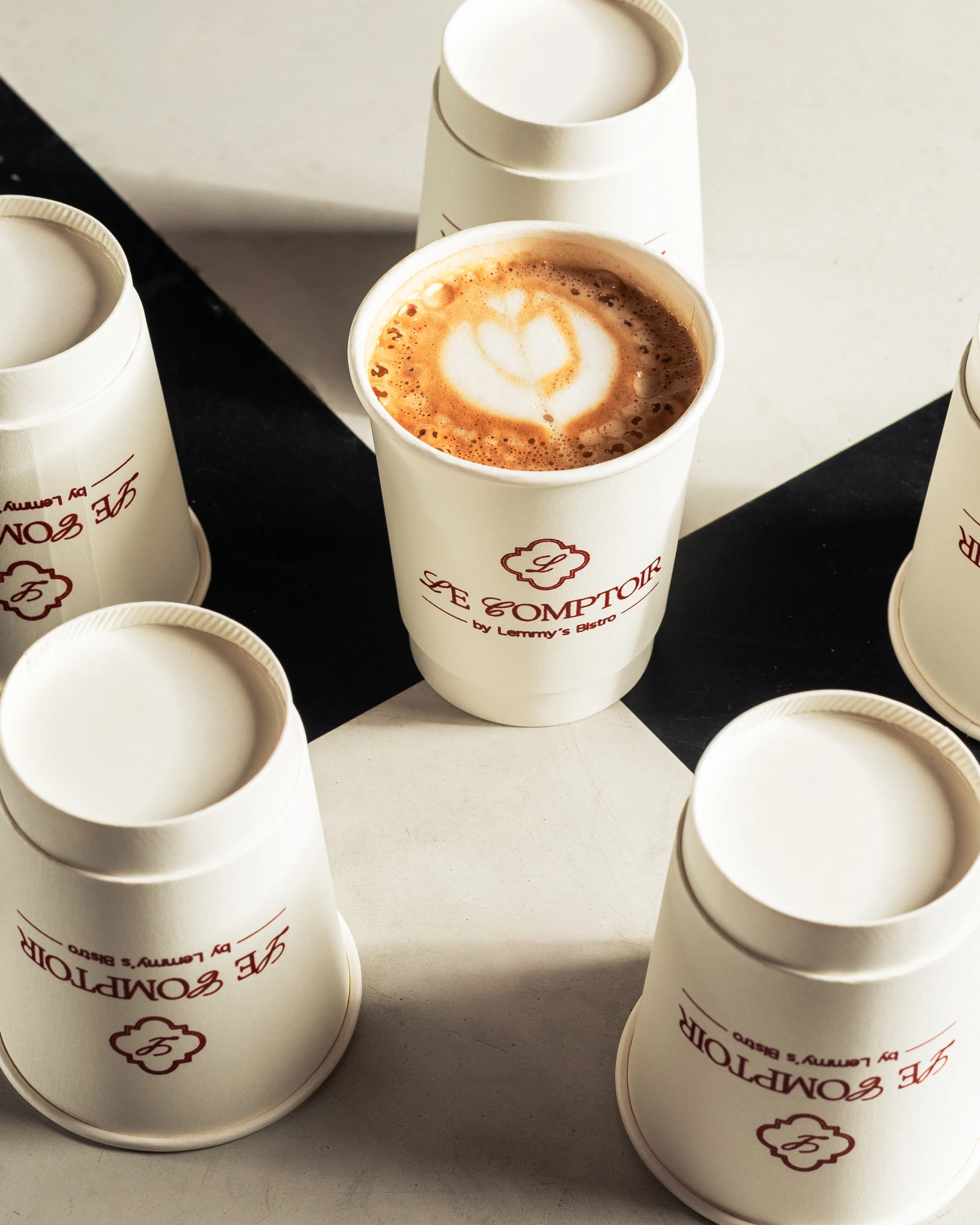 A paper cup of hot chocolate with latte art, surrounded by empty cups on a black and white checkered surface.