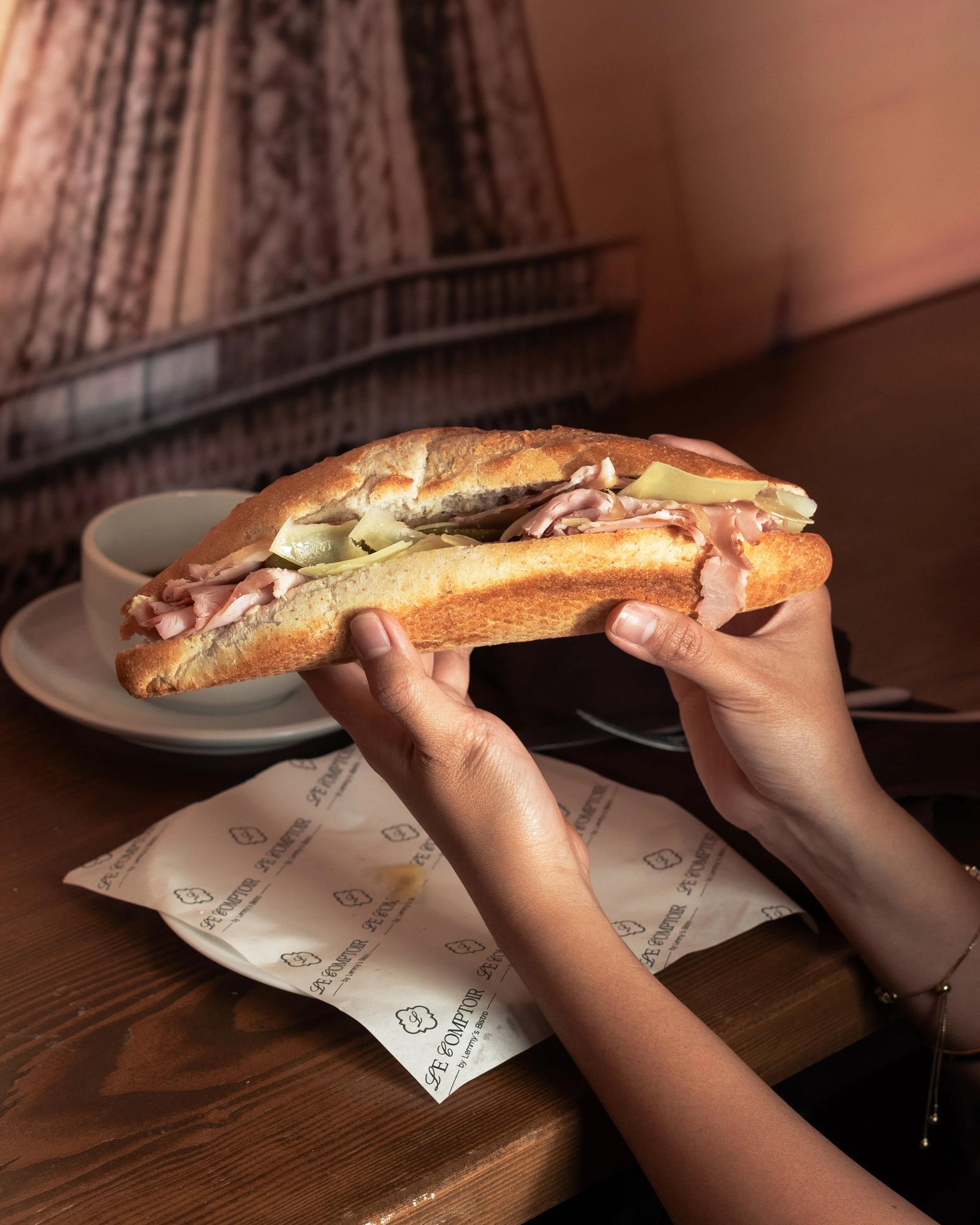 Person holding a sub sandwich with sliced turkey and pickles at a restaurant table, with a small bowl of sauce behind it and a paper with the restaurant's logo on the table.