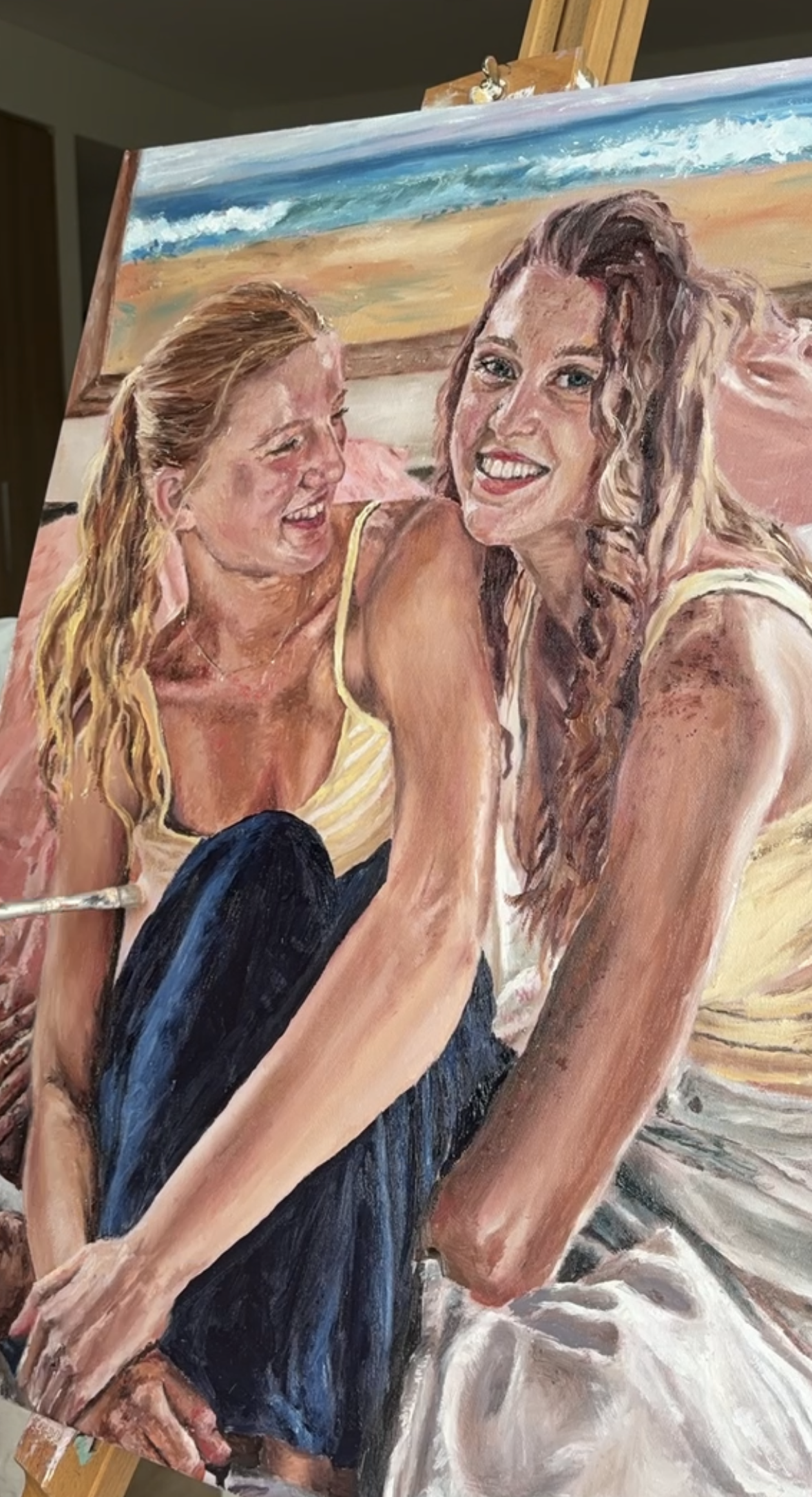A painting of two women smiling and laughing on a beach with the ocean and sky in the background.