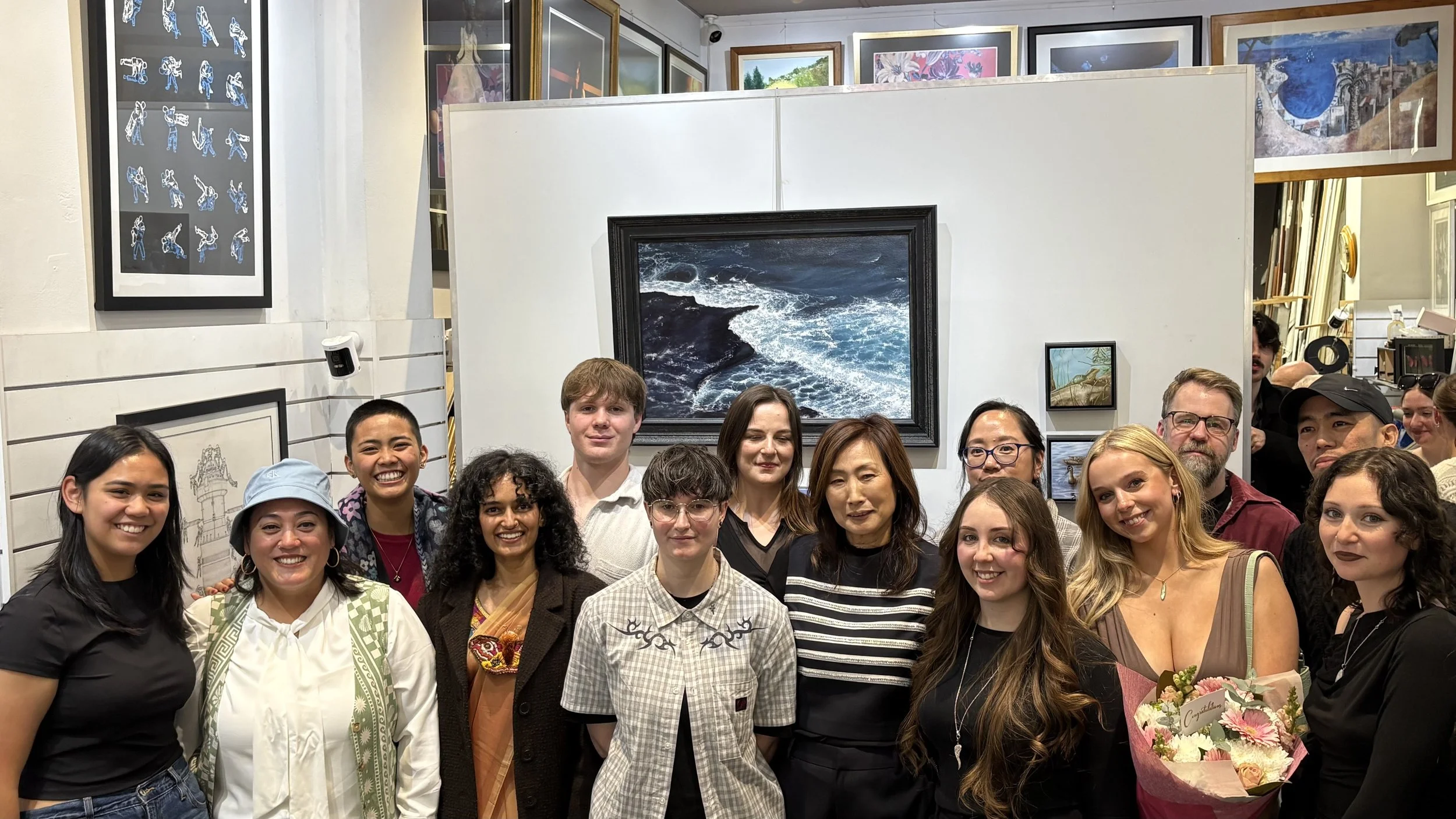 A diverse group of people gathered together at an art gallery, smiling for the photo with artwork displayed on the white walls behind them. Stella's Gallery, Penrith, 2750. New South Wales, Australia.