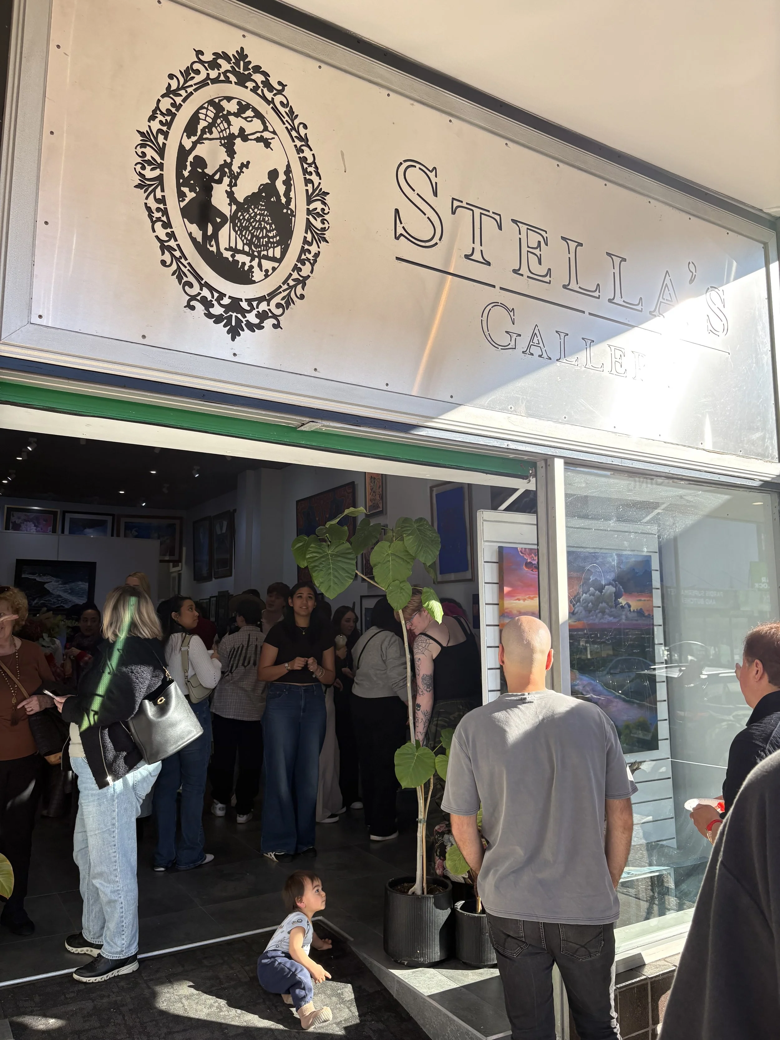 A crowd of people gathered inside an art gallery, with some looking at paintings on the walls and others waiting in line.. Stella's gallery, in Penrith, Sydney, New South Wales, Australia.