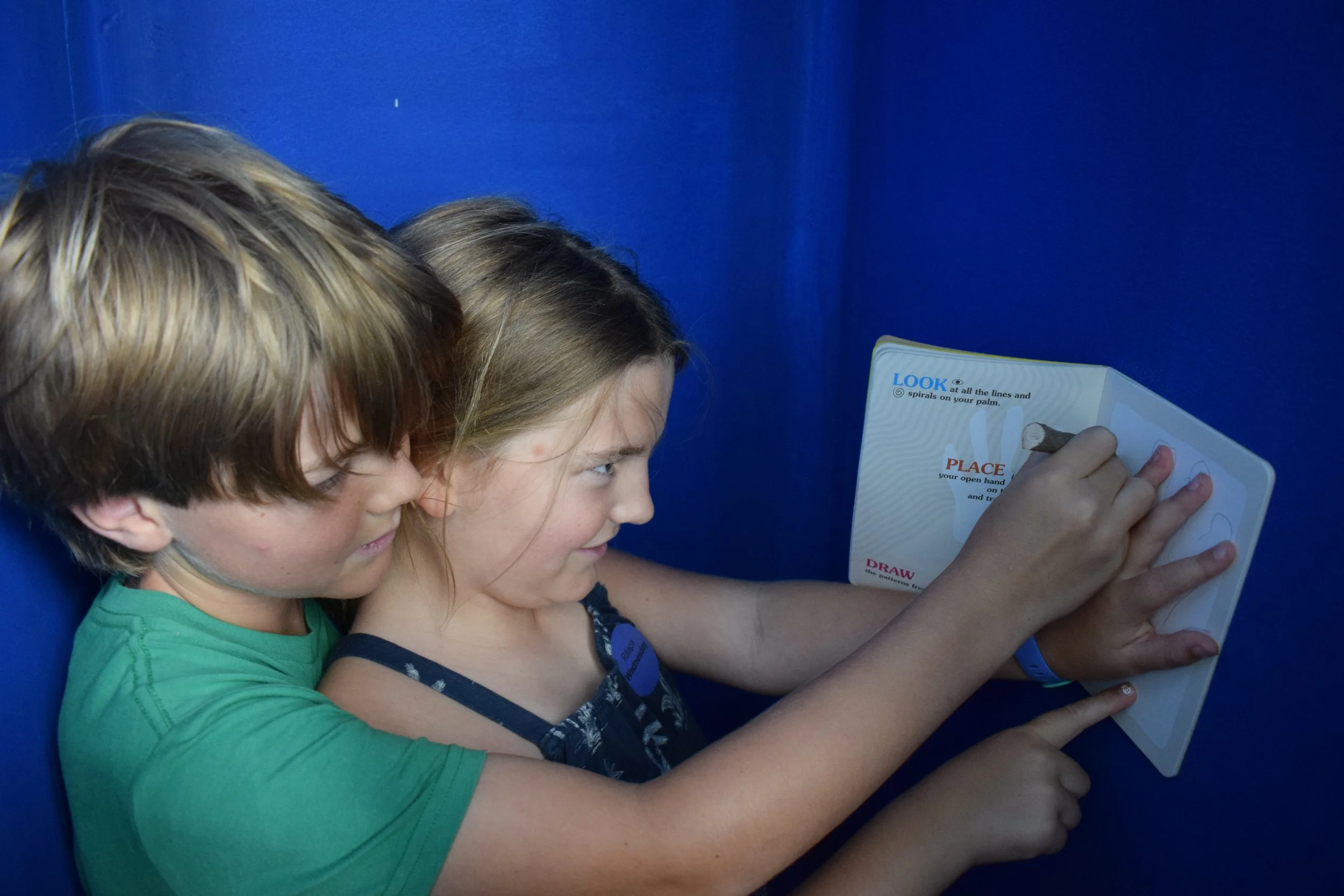 Two children, a boy and a girl, closely reading a booklet with instructions and illustrations while sitting against a blue wall.