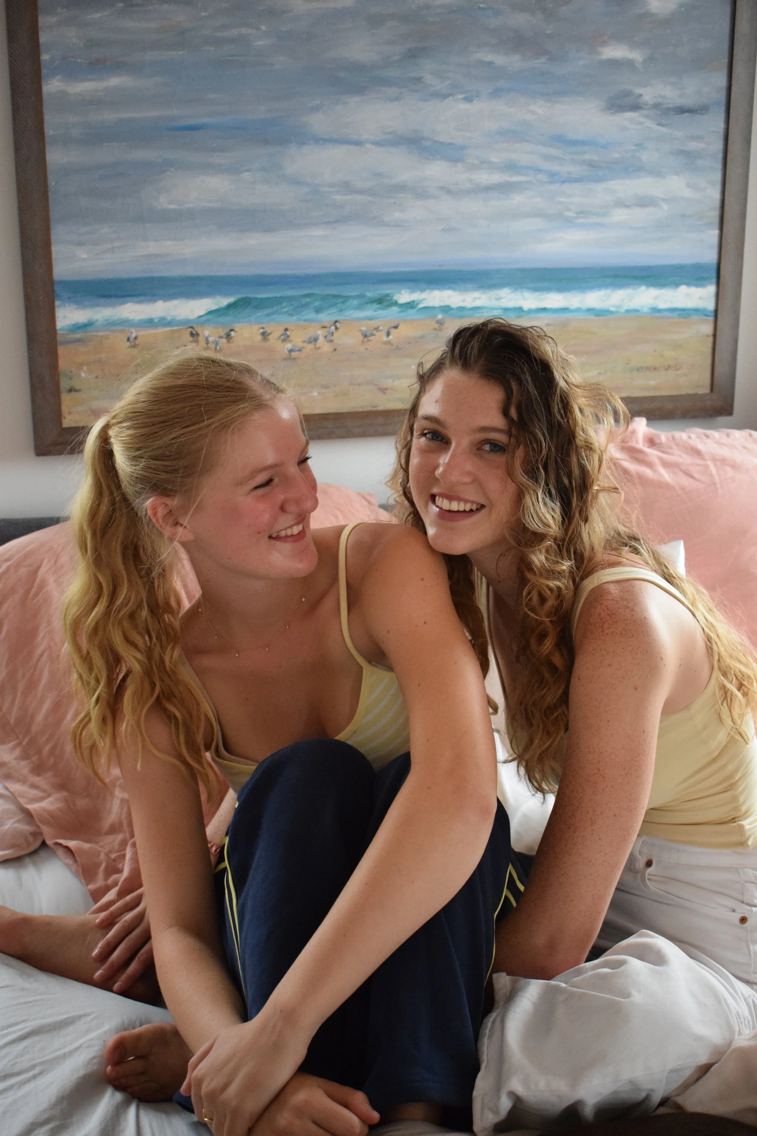 Two young women with long, curly blonde hair smiling and sitting close together on a bed. One is wearing a yellow and white striped tank top, the other a yellow tank top. Behind them, there is a large painting of a beach scene with waves and seagulls.