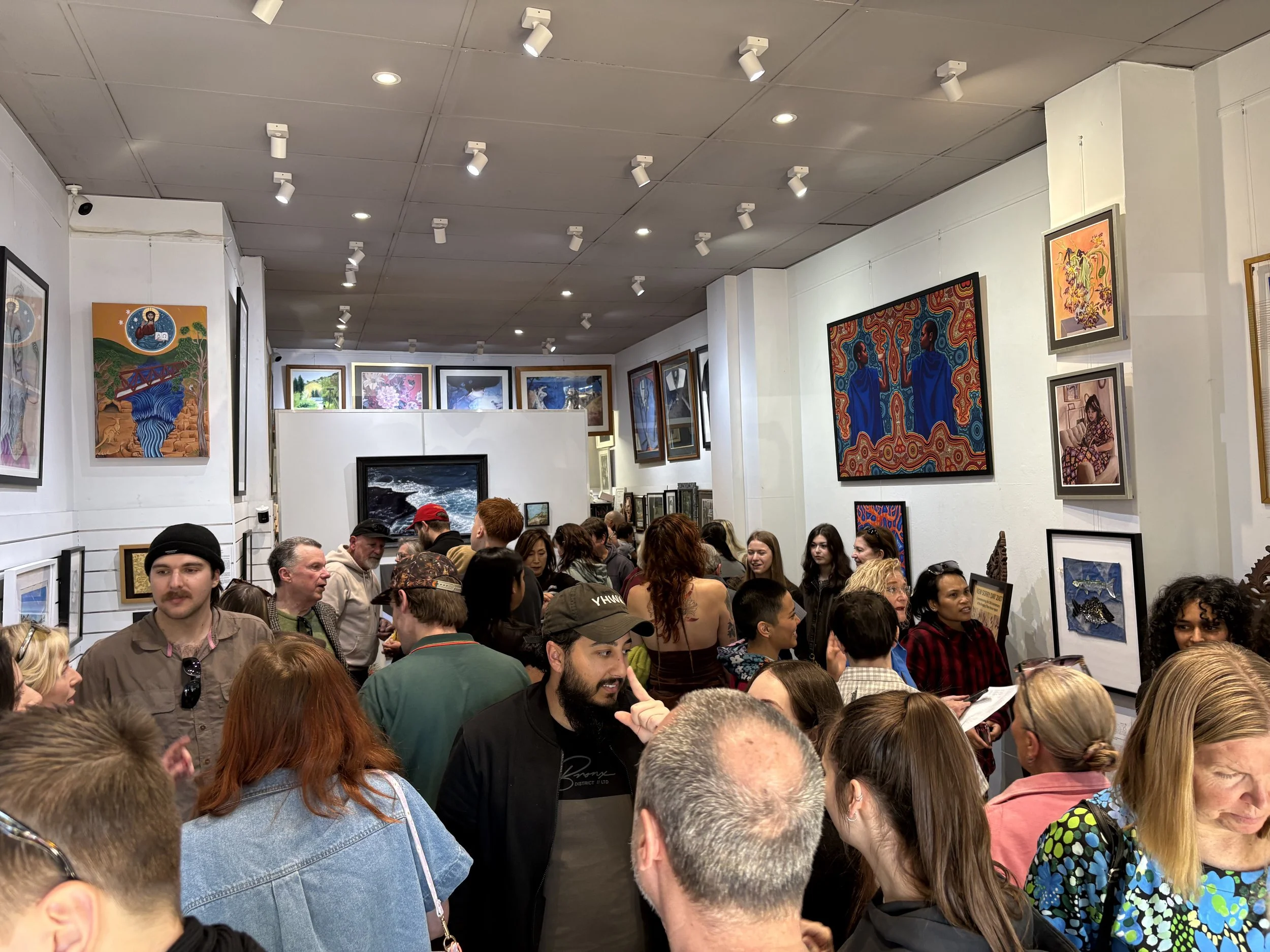 Crowd of people inside an art gallery, looking at various artworks displayed on white walls and in frames. Stella's Gallery, Penrith, Australia.