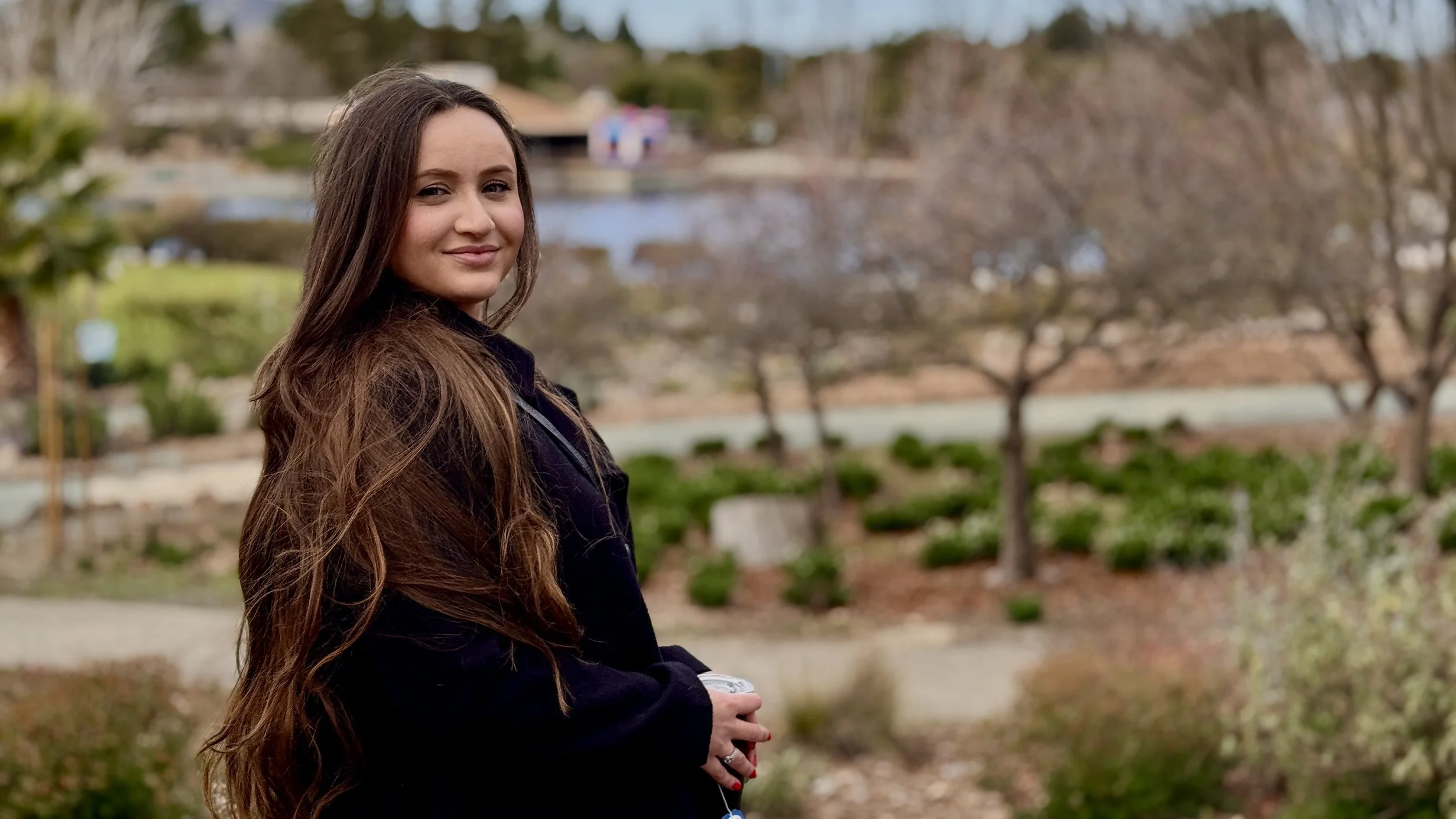 Young woman with long brown hair wearing a dark coat standing outdoors with trees and a pond in the background.