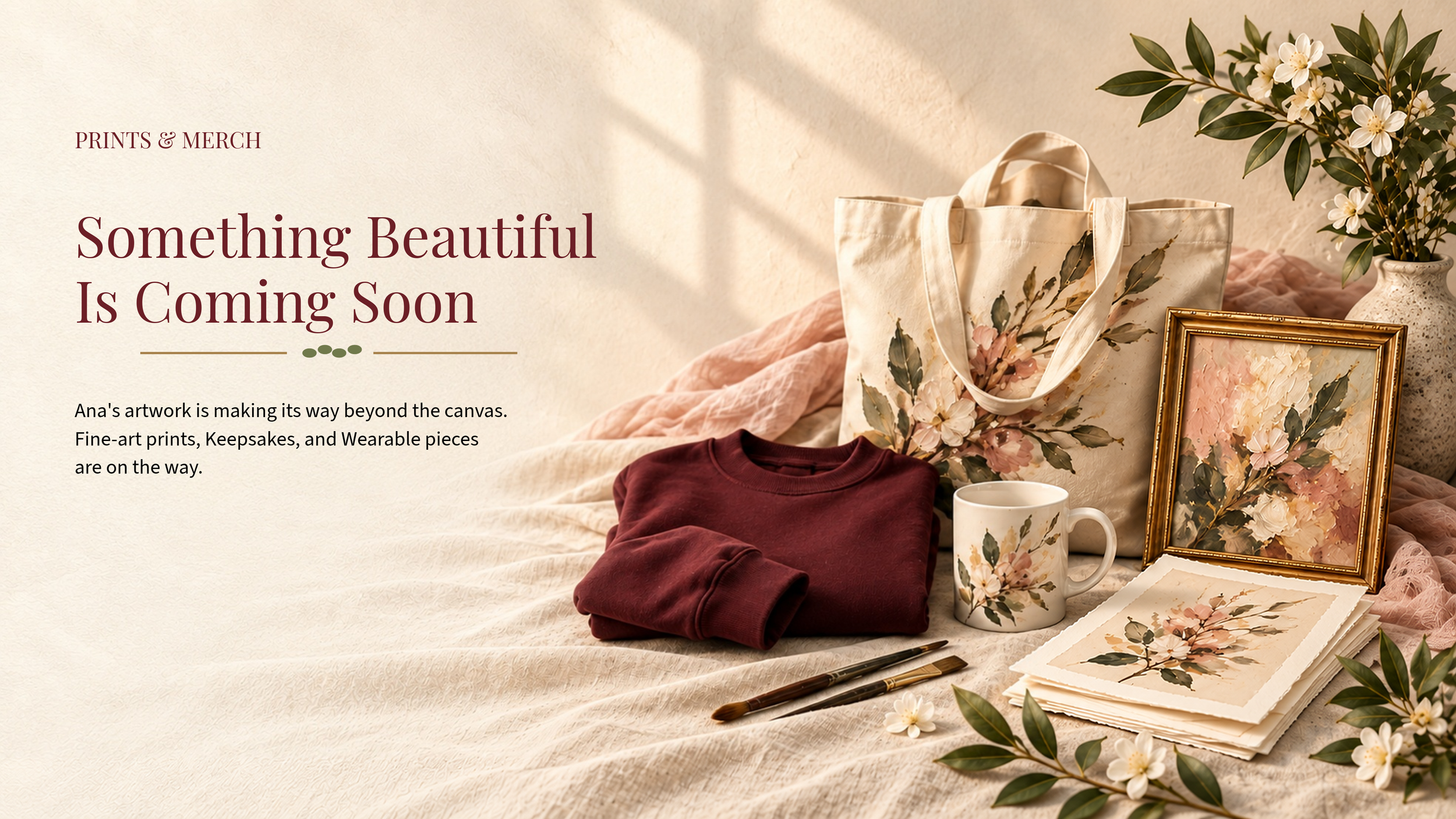 A website banner with a beige background and dark red text that says "Something Beautiful Is Coming Soon" and promotes prints and merchandise by Ana. The image features a flat lay arrangement of a maroon sweatshirt, a botanical-themed tote bag, a coffee mug, paint brushes, a framed floral painting, a stack of floral prints, and a vase with white flowers, all on a soft pink fabric, with sunlight and green leaves adding a natural touch.