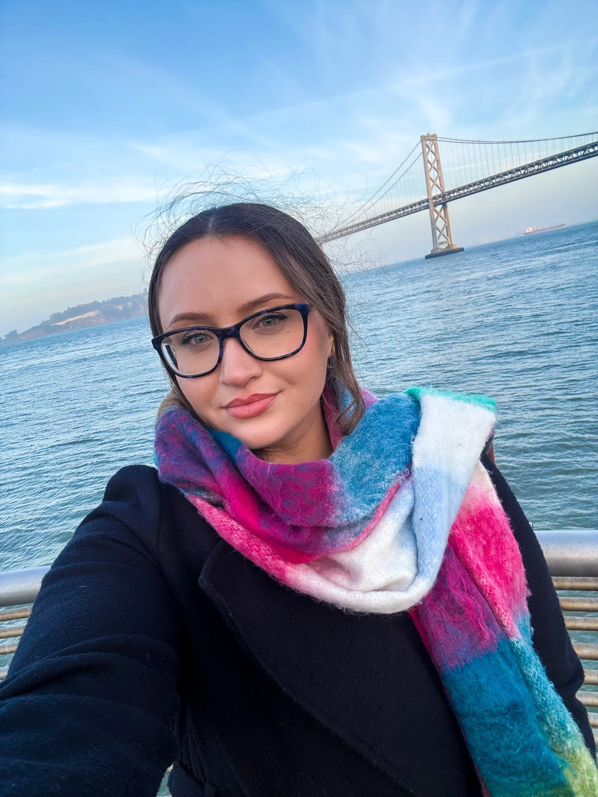 A woman with glasses and a colorful scarf taking a selfie on a boat with the San Francisco Bay Bridge in the background.