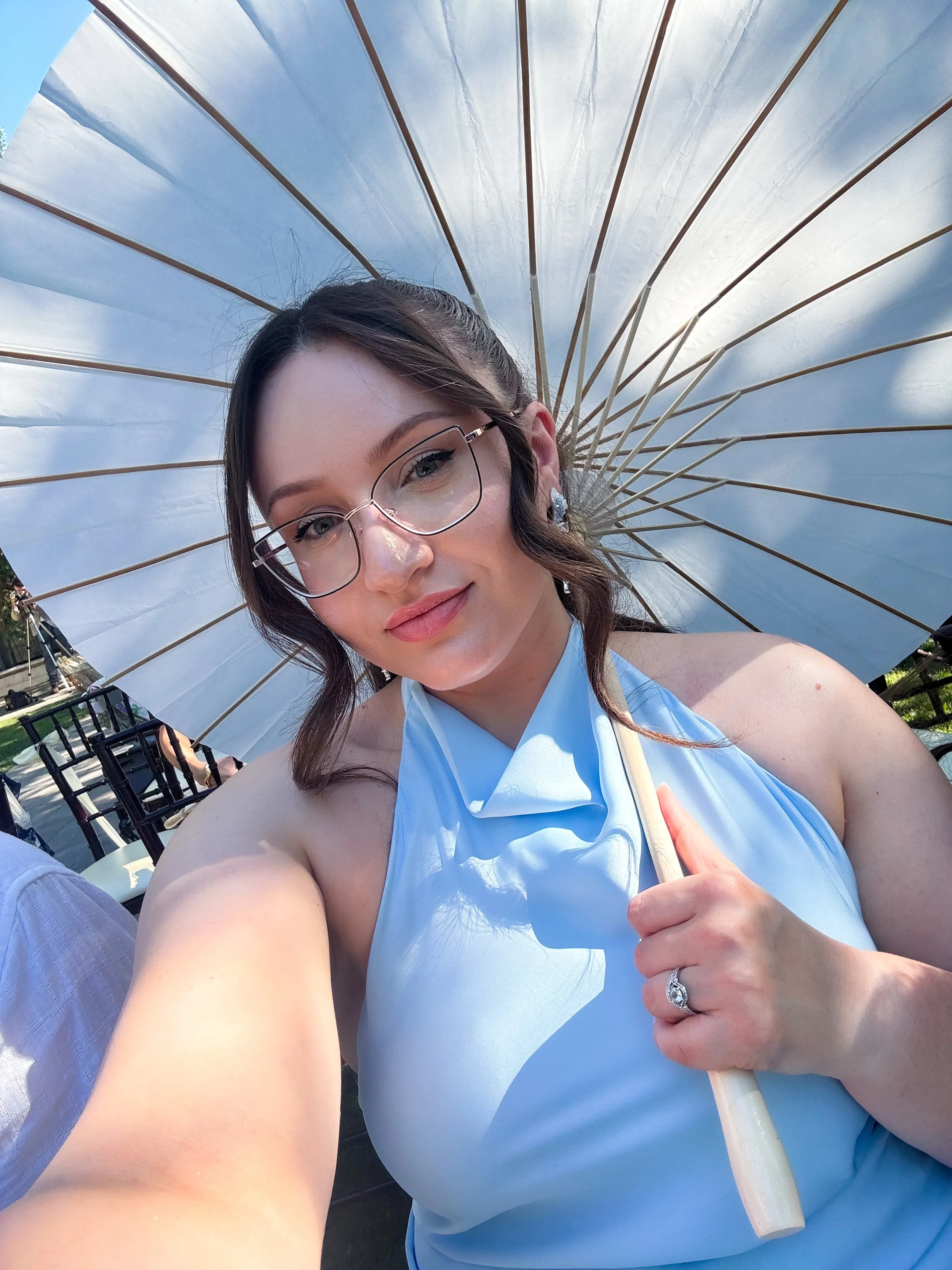 A woman posing for a selfie under a large white parasol, wearing glasses and a light blue halter dress, holding the parasol handle with her right hand.