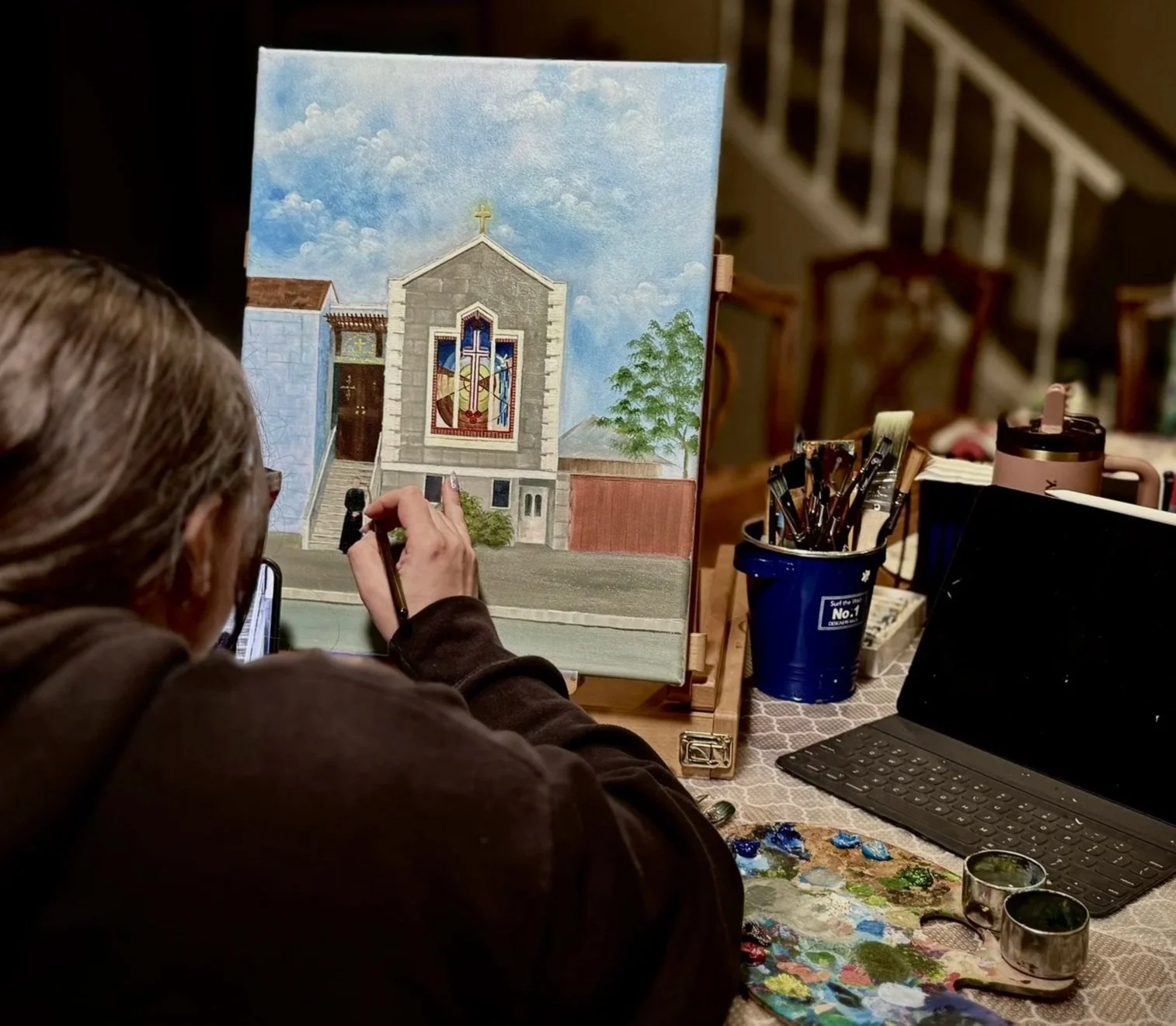 An artist painting a church on a canvas, with painting supplies and a laptop on a table.